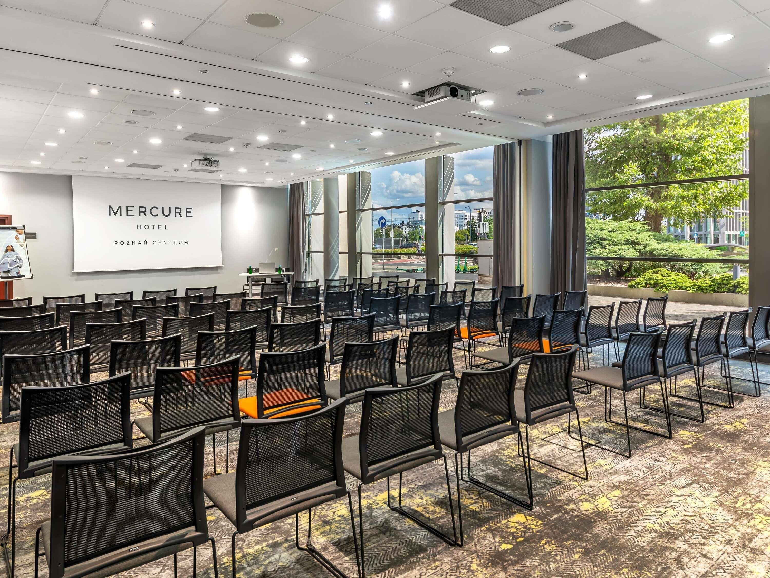 A conference room with rows of chairs, a projector screen, and large windows offering a view of greenery and buildings.