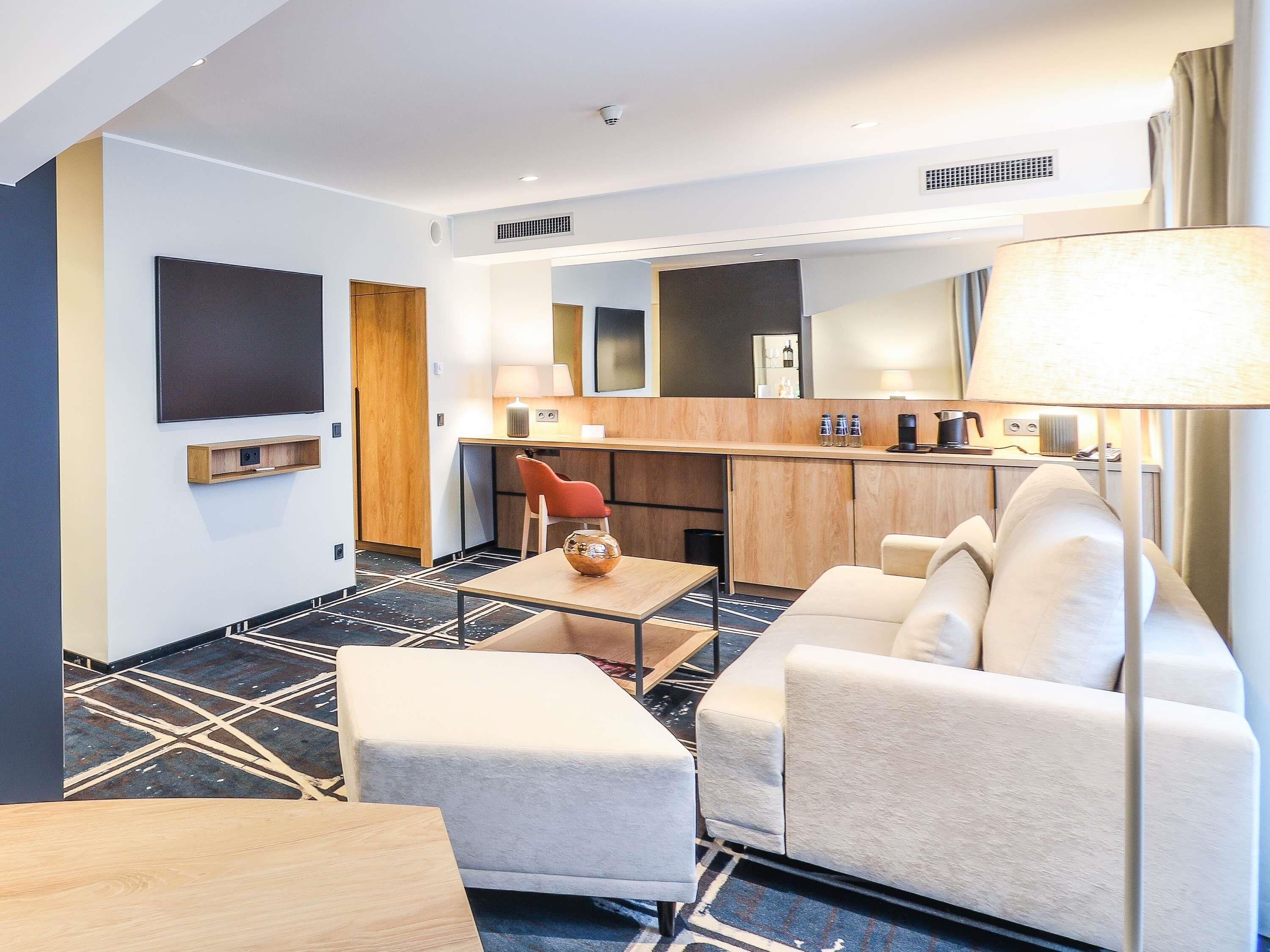A modern hotel room with a sofa, a small table, a TV, a wooden desk, and a kitchenette.