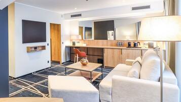 A modern hotel room with a sofa, a small table, a TV, a wooden desk, and a kitchenette.