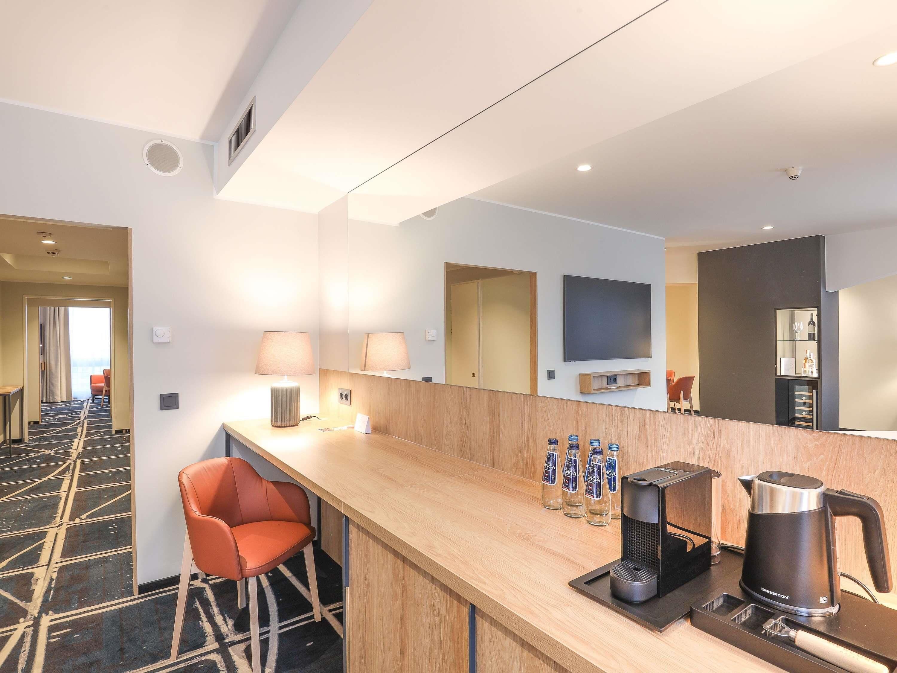 A hotel room with a wooden bar, two chairs, a coffee maker, and a flat-screen TV mounted on the wall.