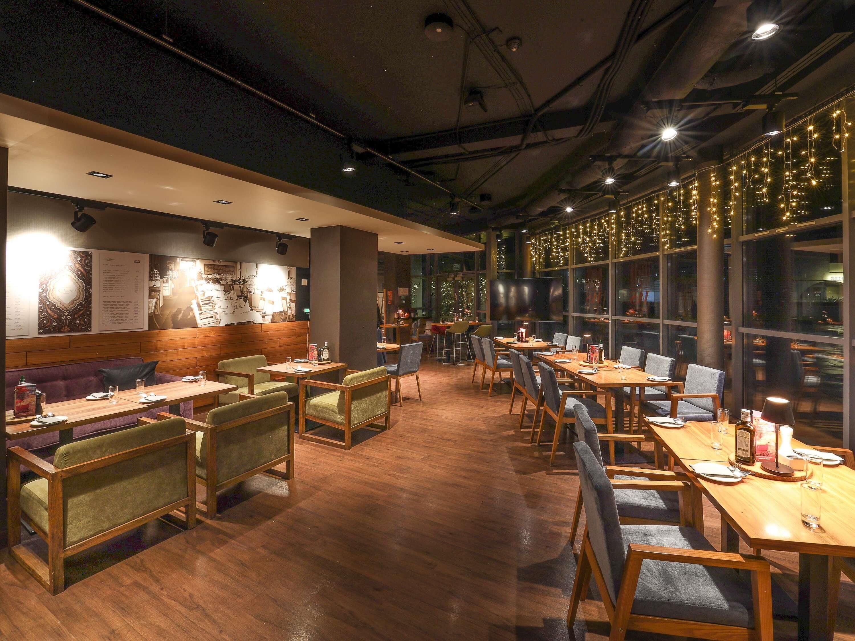 A restaurant with wooden tables and chairs, a menu board, and a large window with string lights.