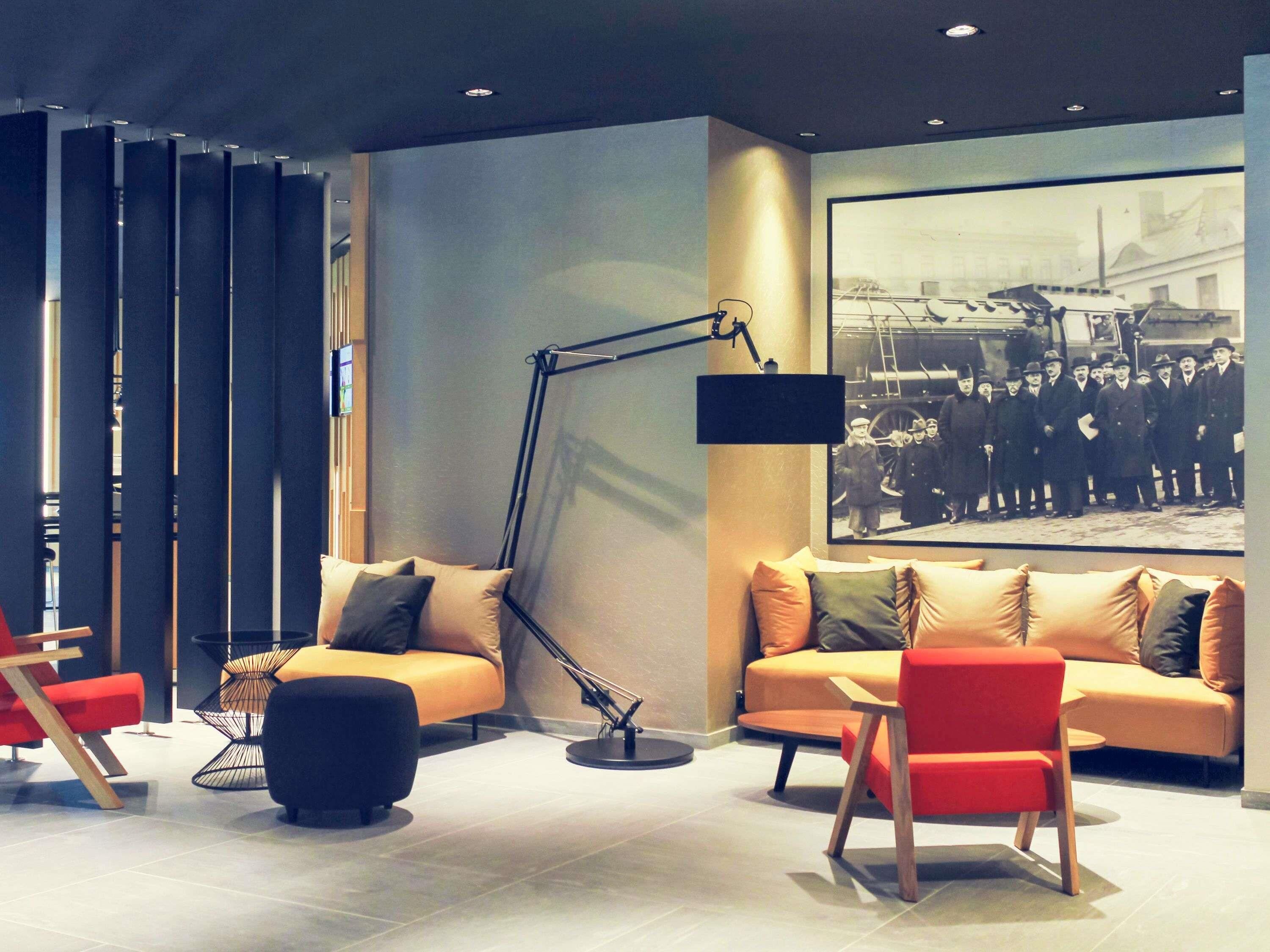 A modern hotel lobby with a sofa, ottoman, and chairs, a large black floor lamp, and a historical photograph on the wall.