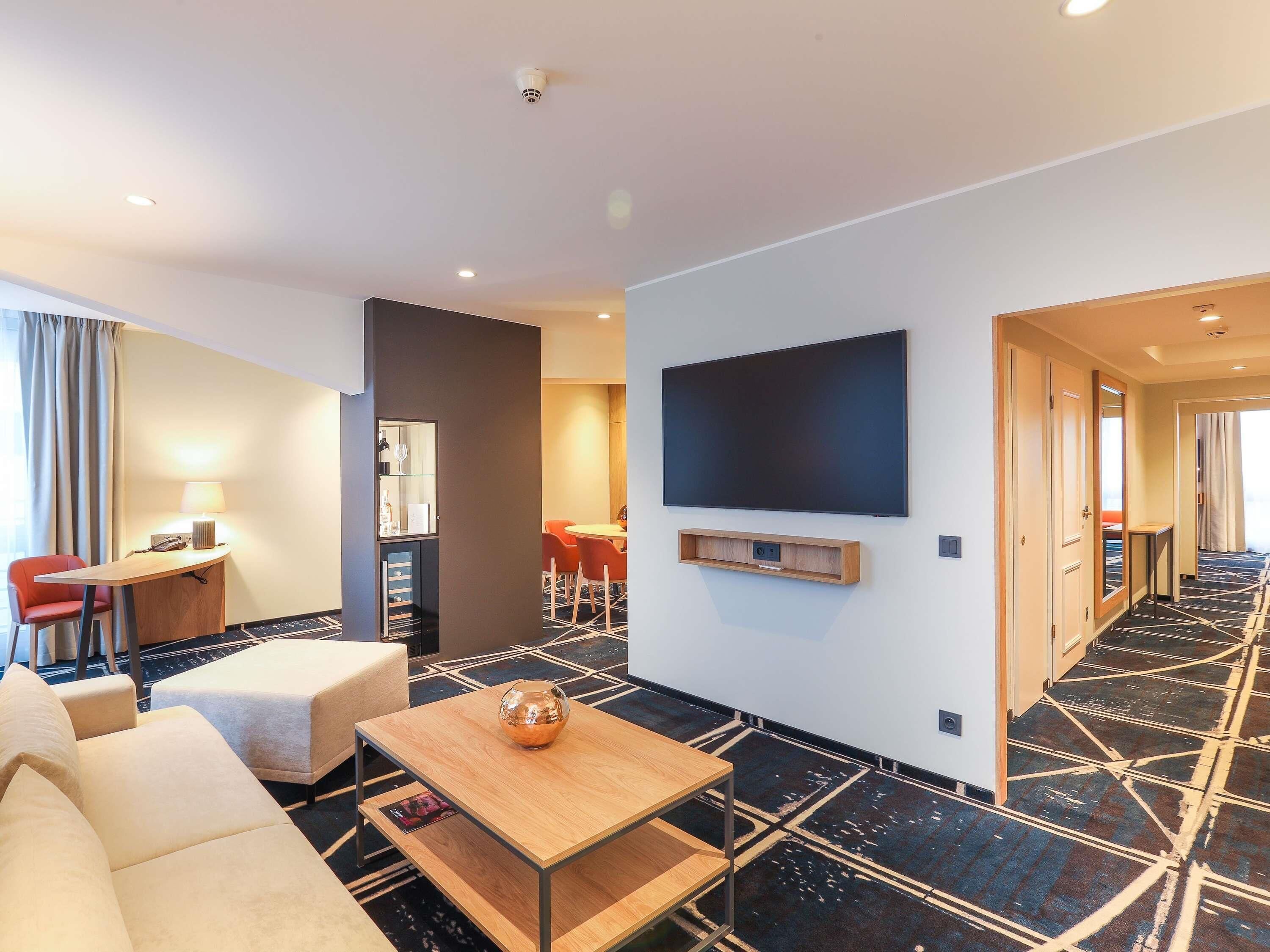 Une chambre d’hôtel moderne équipée d’un canapé, d’une table basse, d’un téléviseur fixé au mur, d’un bureau avec une chaise et d’un coin repas avec des chaises.