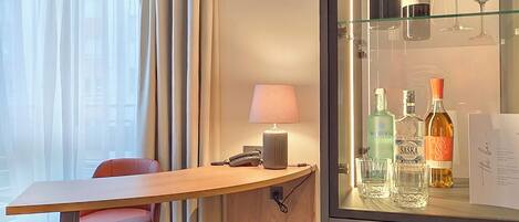 A hotel room with a wooden desk, a red chair, a lamp, and a minibar with bottles and glasses.