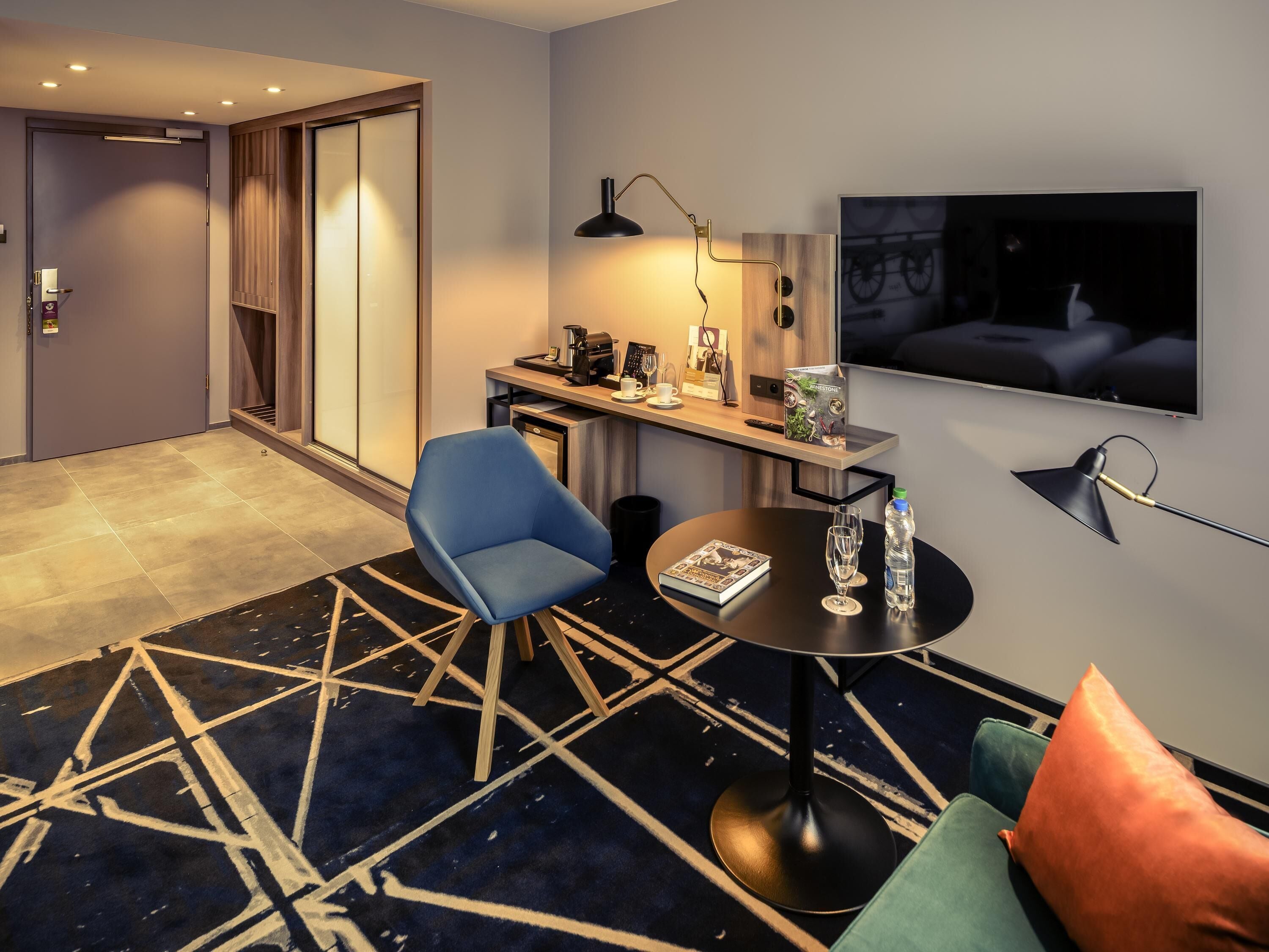 A hotel room with a desk, chair, TV, and a patterned rug.