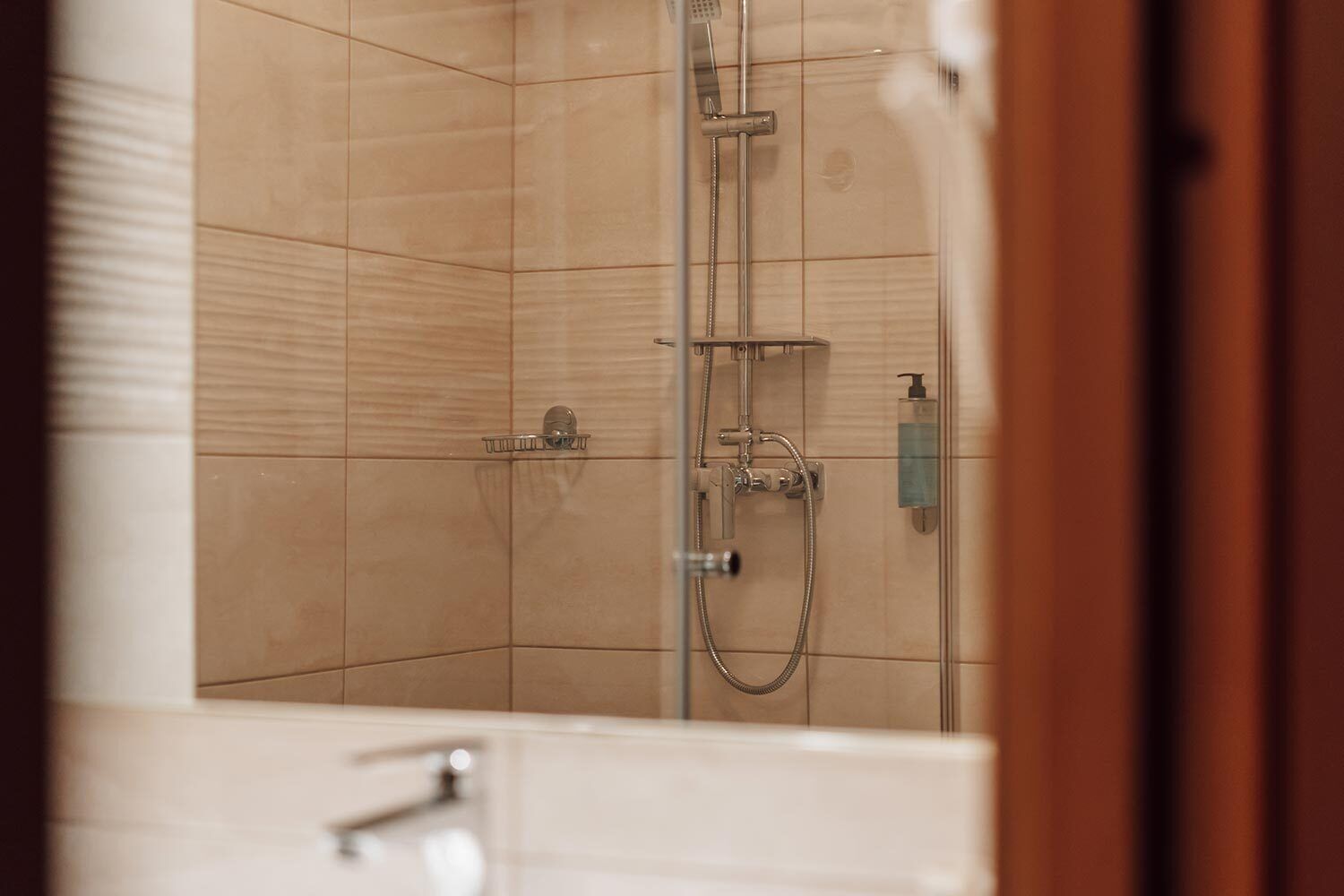 A shower area with tiled walls, a handheld showerhead, a wall-mounted faucet, and a soap dispenser.