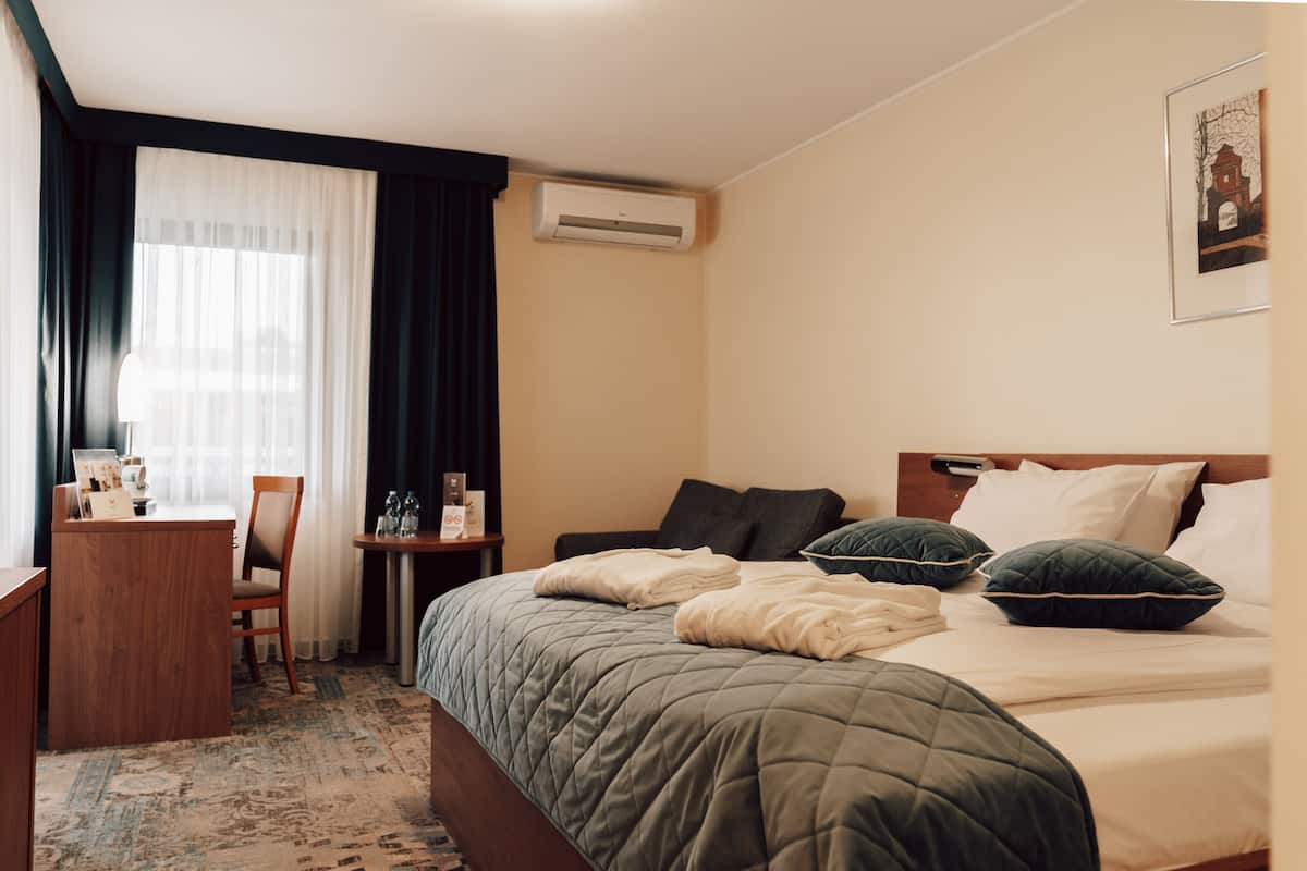 A hotel room with a bed, a desk, a chair, a small table, and an air conditioning unit.
