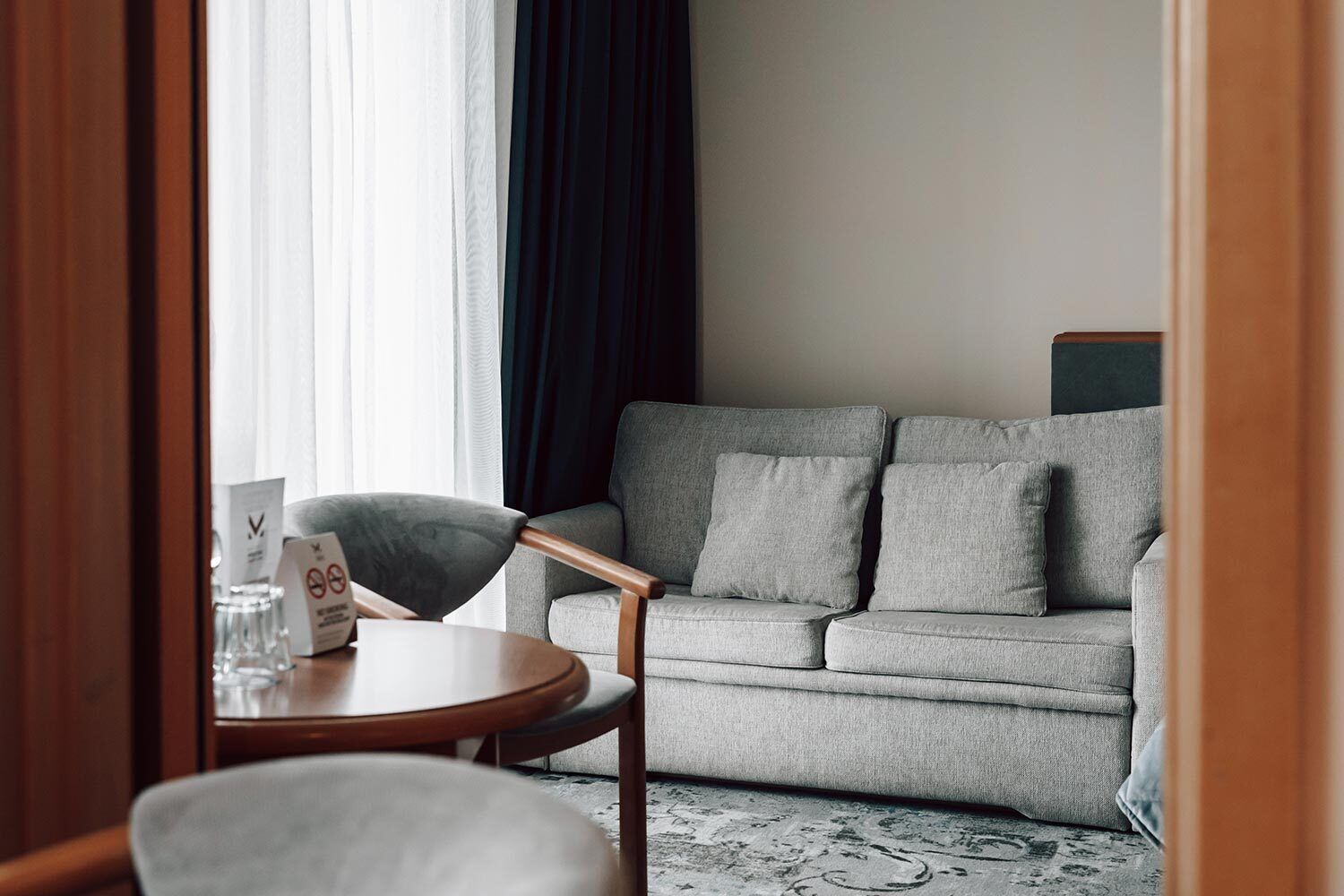 Una habitación de hotel moderna con un sofá gris, una silla de madera y una pequeña mesa redonda.