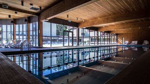 A modern indoor swimming pool with clear glass walls, wooden beams, and sun loungers.