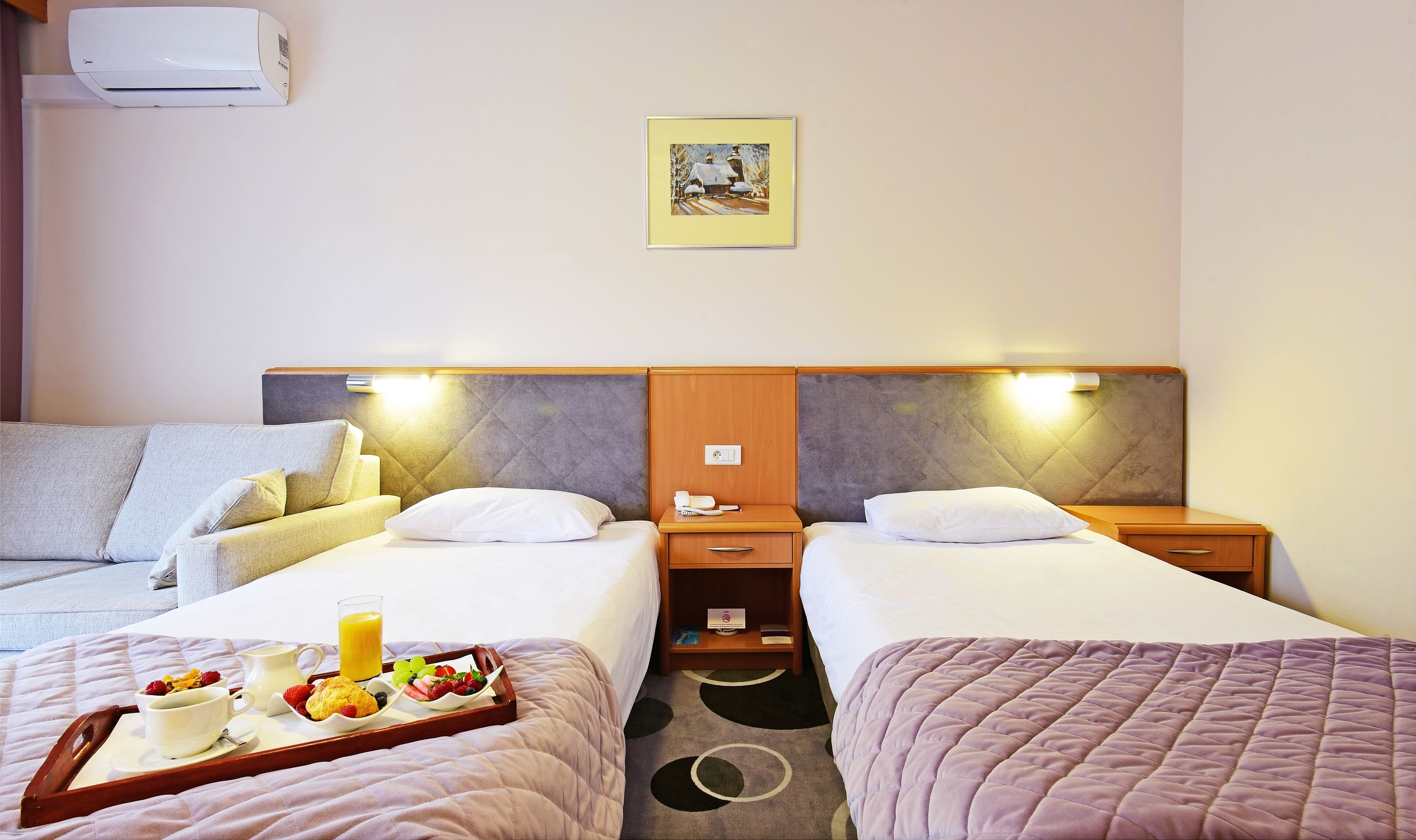 Habitación de hotel con dos camas, una mesita de noche con lámpara, aire acondicionado de pared y un cuadro enmarcado.