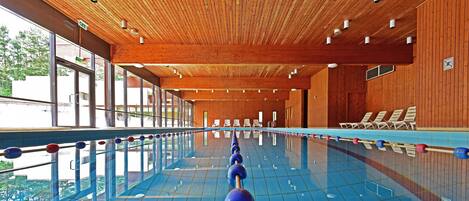 An indoor swimming pool with wooden paneling, clear glass windows, and lounge chairs along the side.