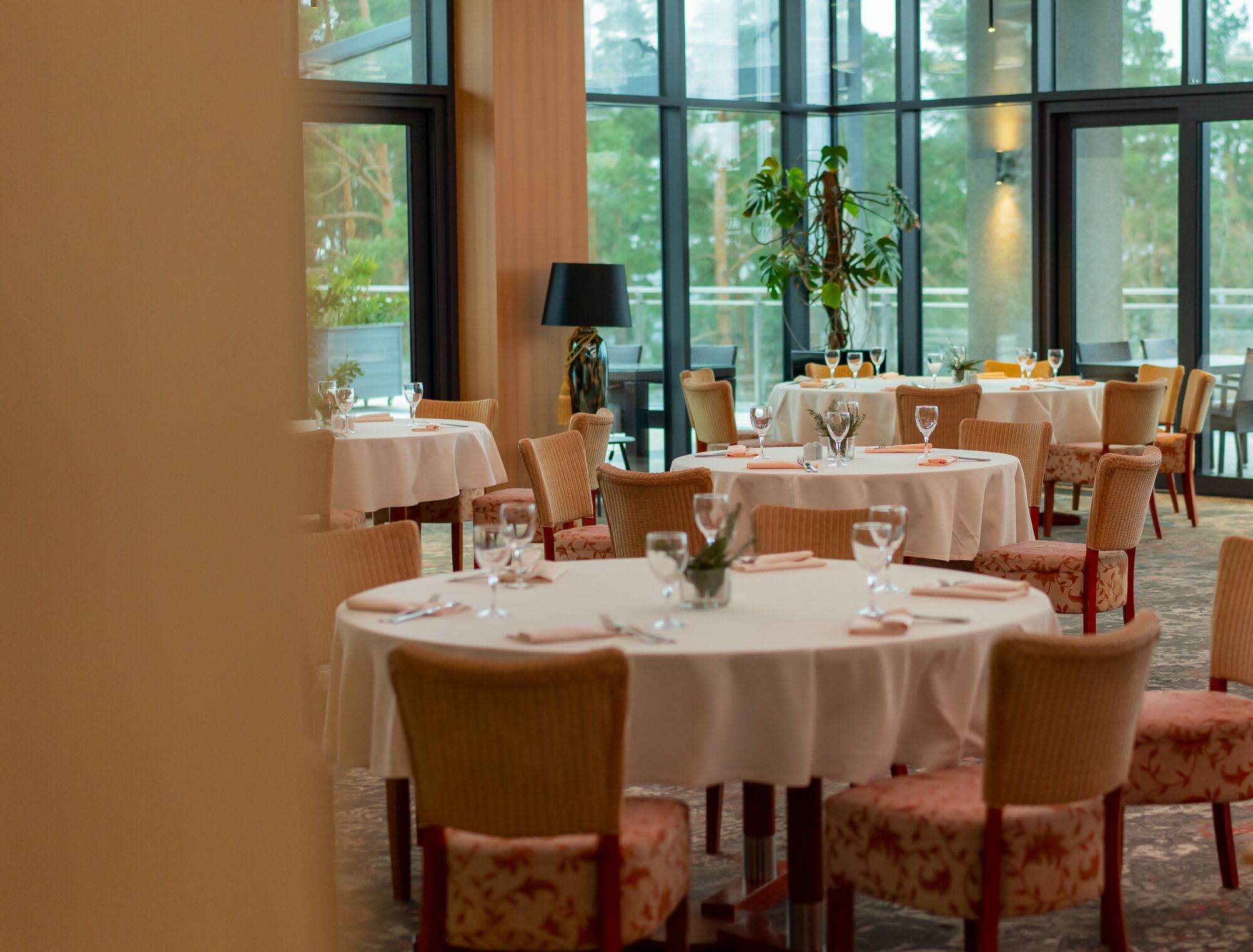 Un restaurante con mesas redondas, sillas acolchadas, ventanas amplias y plantas de interior.