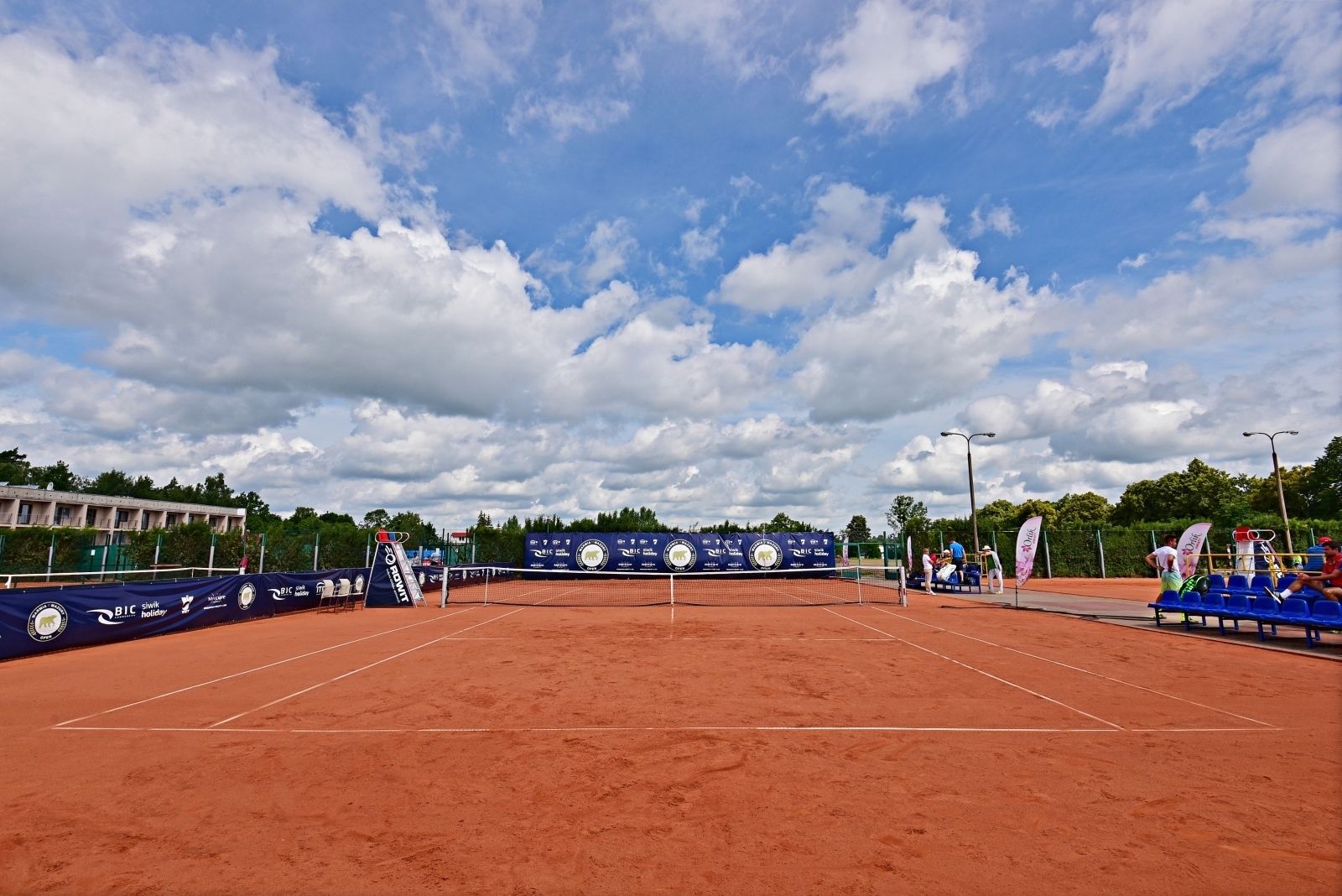 Un court de tennis en terre battue avec un filet au centre, entouré de sièges bleus et de banderoles.