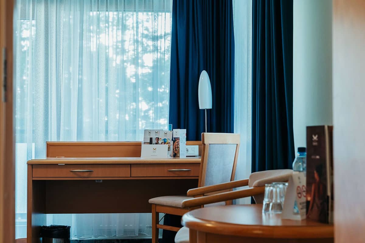 A hotel room with a wooden desk, a chair, a small table, and a window with sheer curtains.