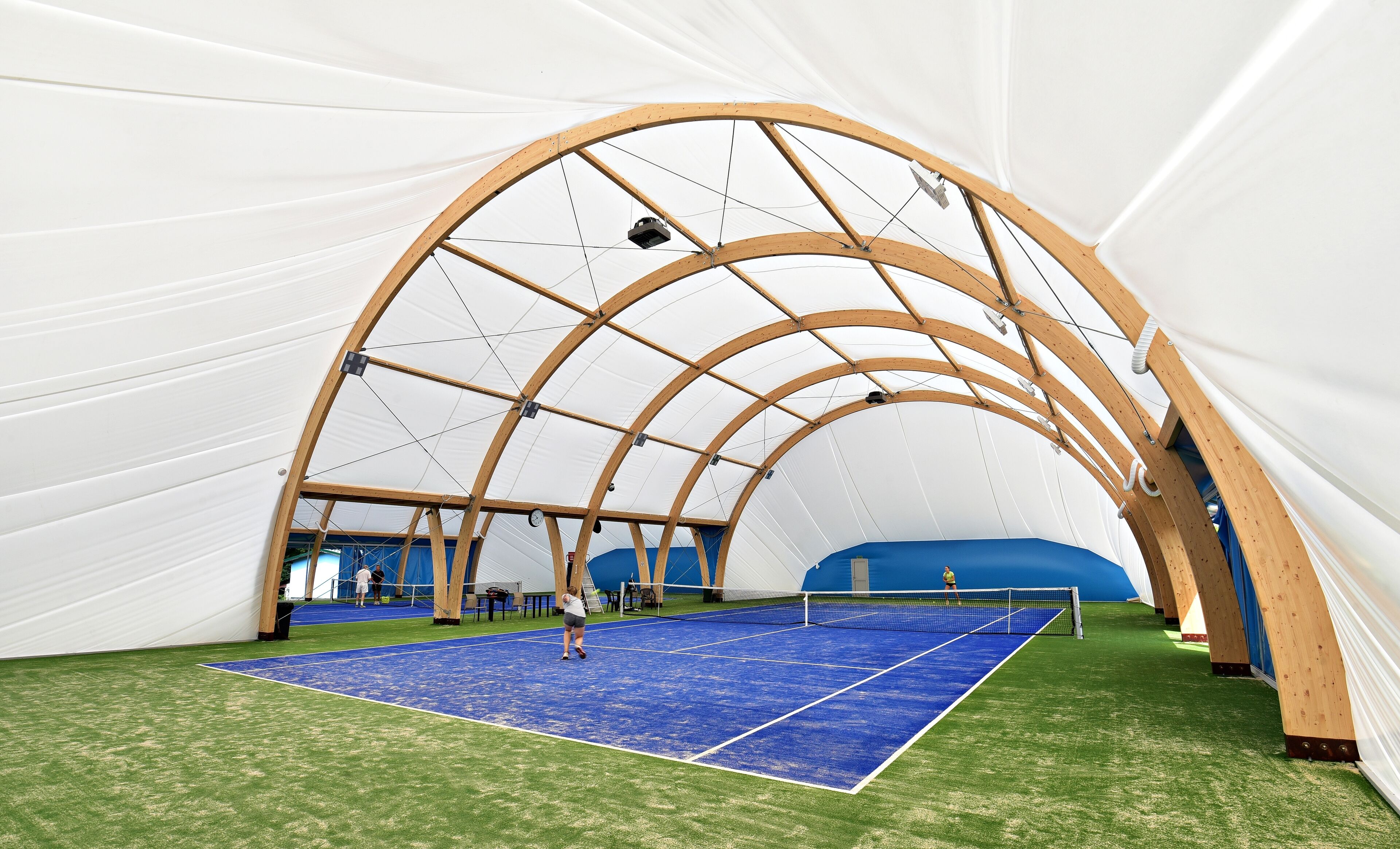 An indoor tennis court with a wooden arch structure, artificial turf, and a blue playing surface.