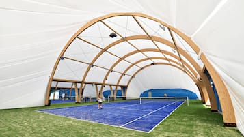 An indoor tennis court with a wooden arch structure, artificial turf, and a blue playing surface.