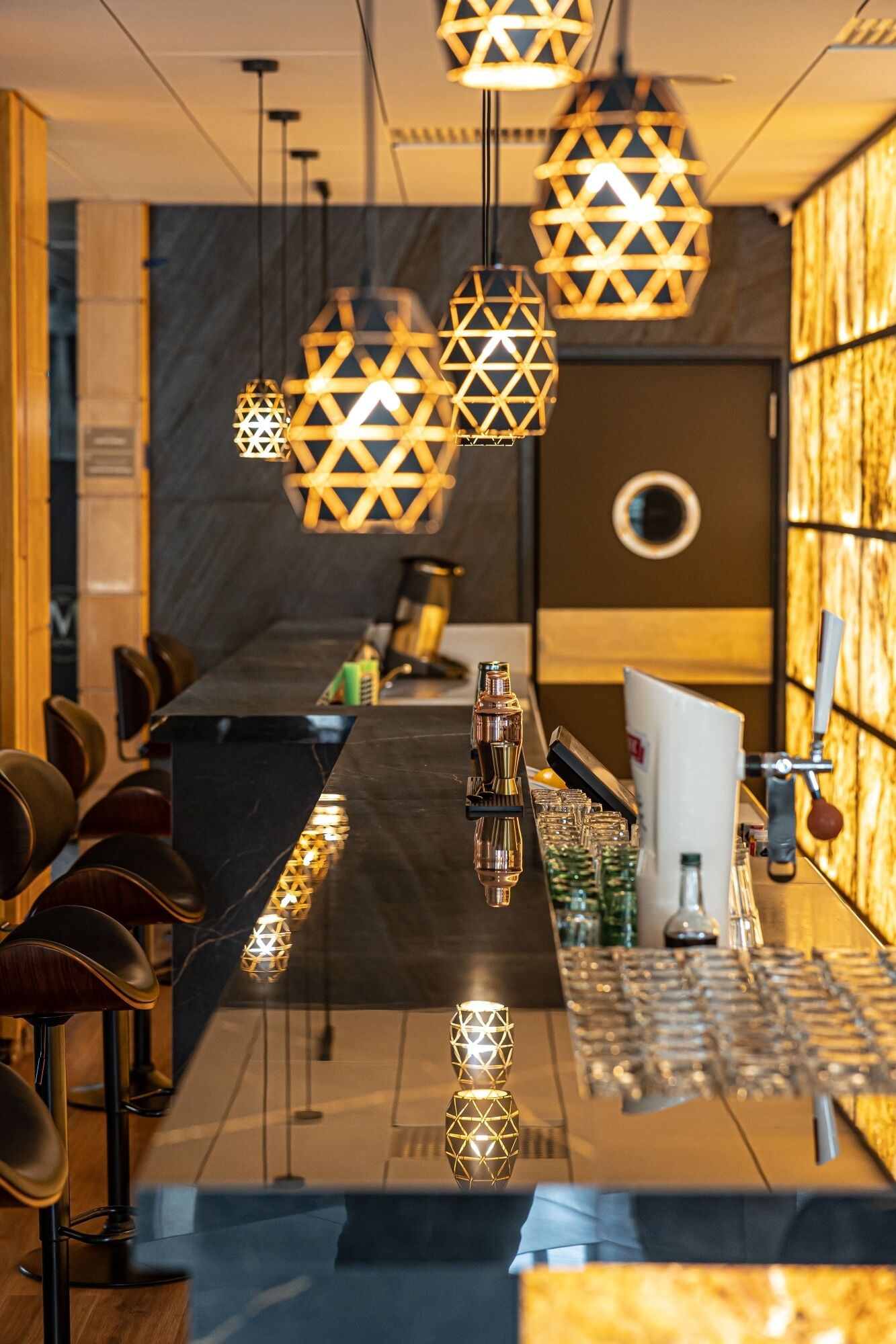 Zona de bar moderna con lámparas colgantes geométricas, una barra larga y taburetes.