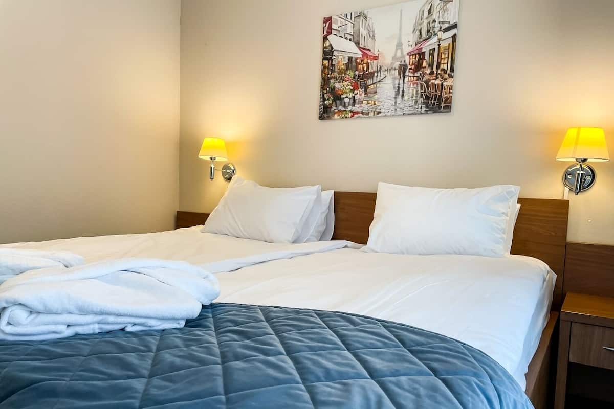 A neatly made bed with a blue quilt, white pillows, and a white blanket folded at the foot. There are two wall-mounted lamps on either side of the bed.