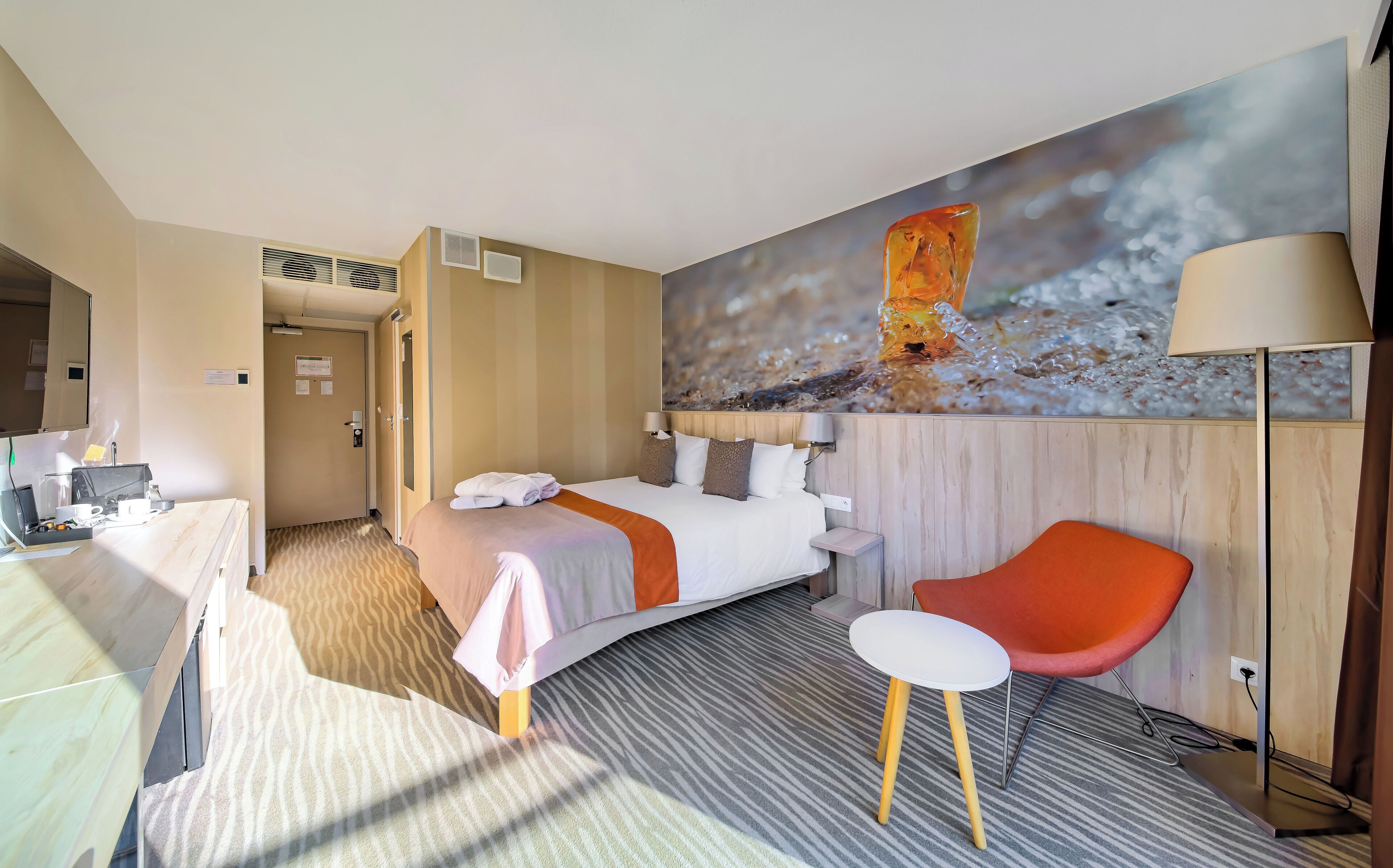 A hotel room with a bed, a desk, a chair, a painting, and a lamp.