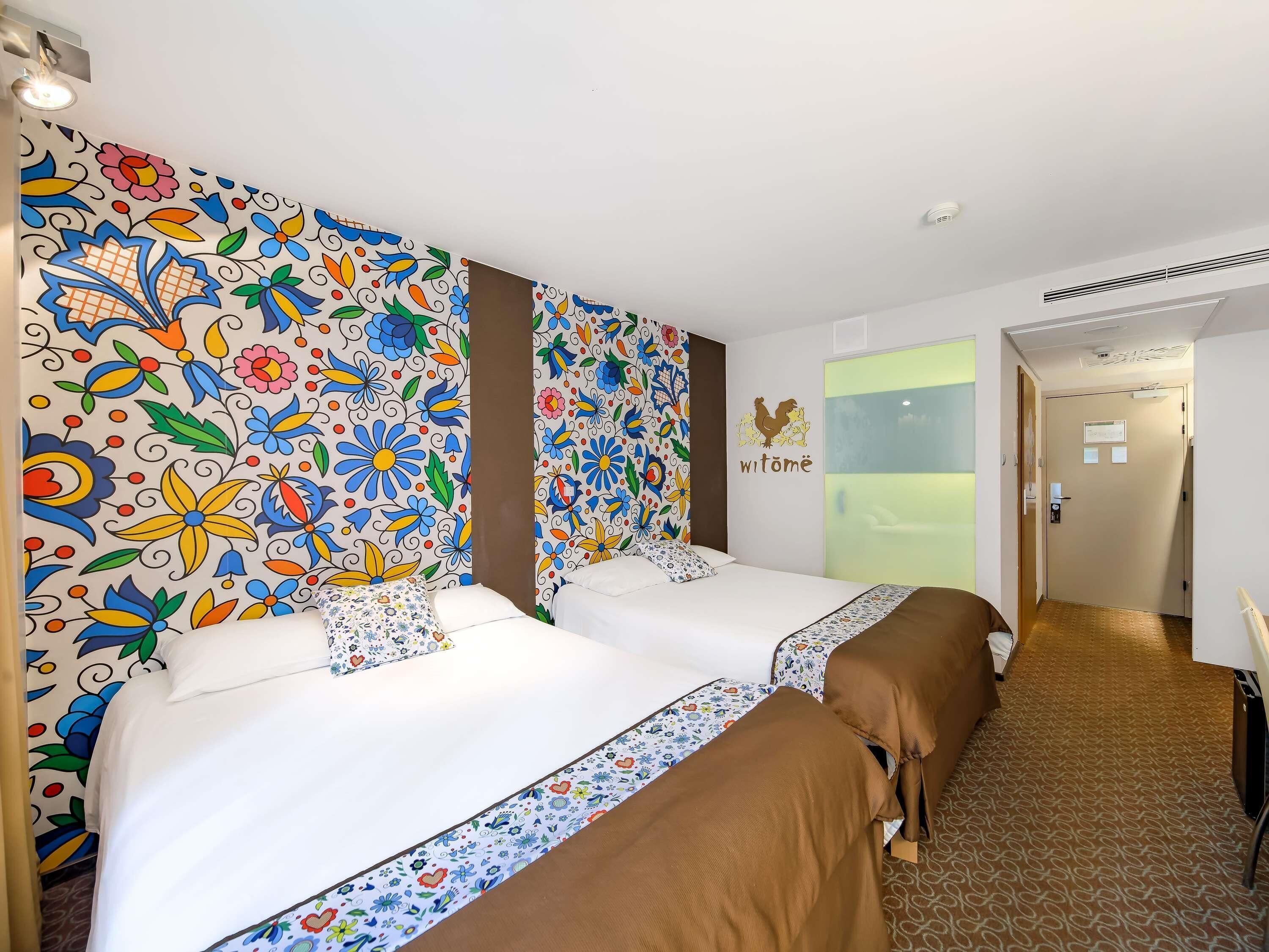 A hotel room with two beds, colorful floral wallpaper, a 'witome' sign, and a view of a green outdoor area.