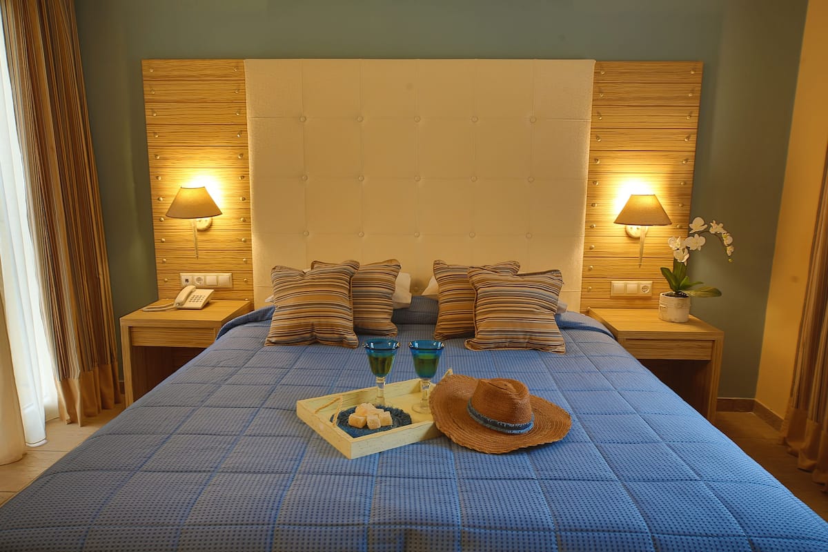 A neatly made bed with a blue checkered bedspread, two bedside lamps, a wooden headboard, and a tray with snacks and a hat on the bed.
