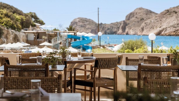 Un área de comedor al aire libre con sillas y mesas de mimbre, con vista a un cuerpo de agua y colinas rocosas.