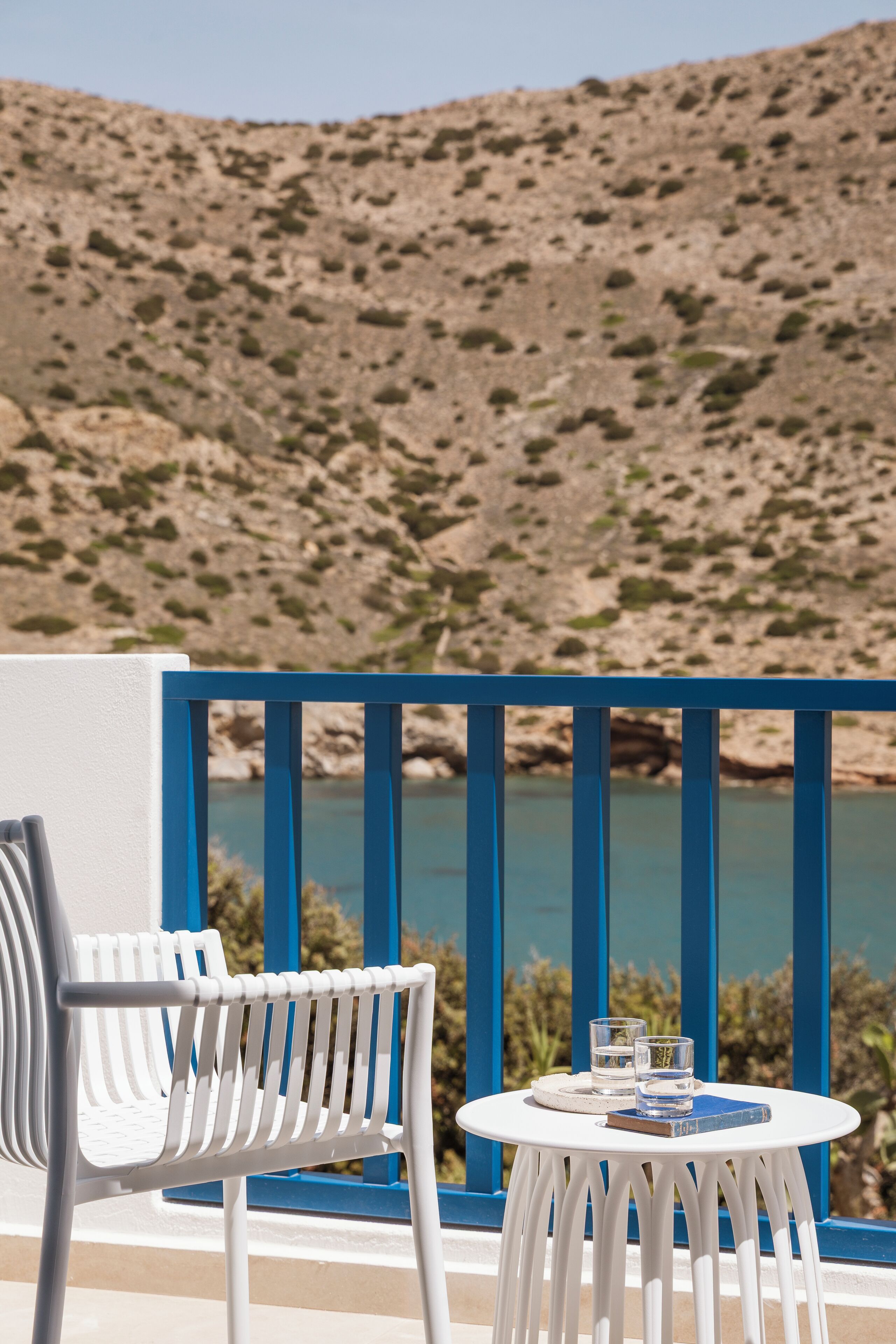 A white outdoor seating area with a table and chairs, overlooking a body of water and a rocky hillside.
