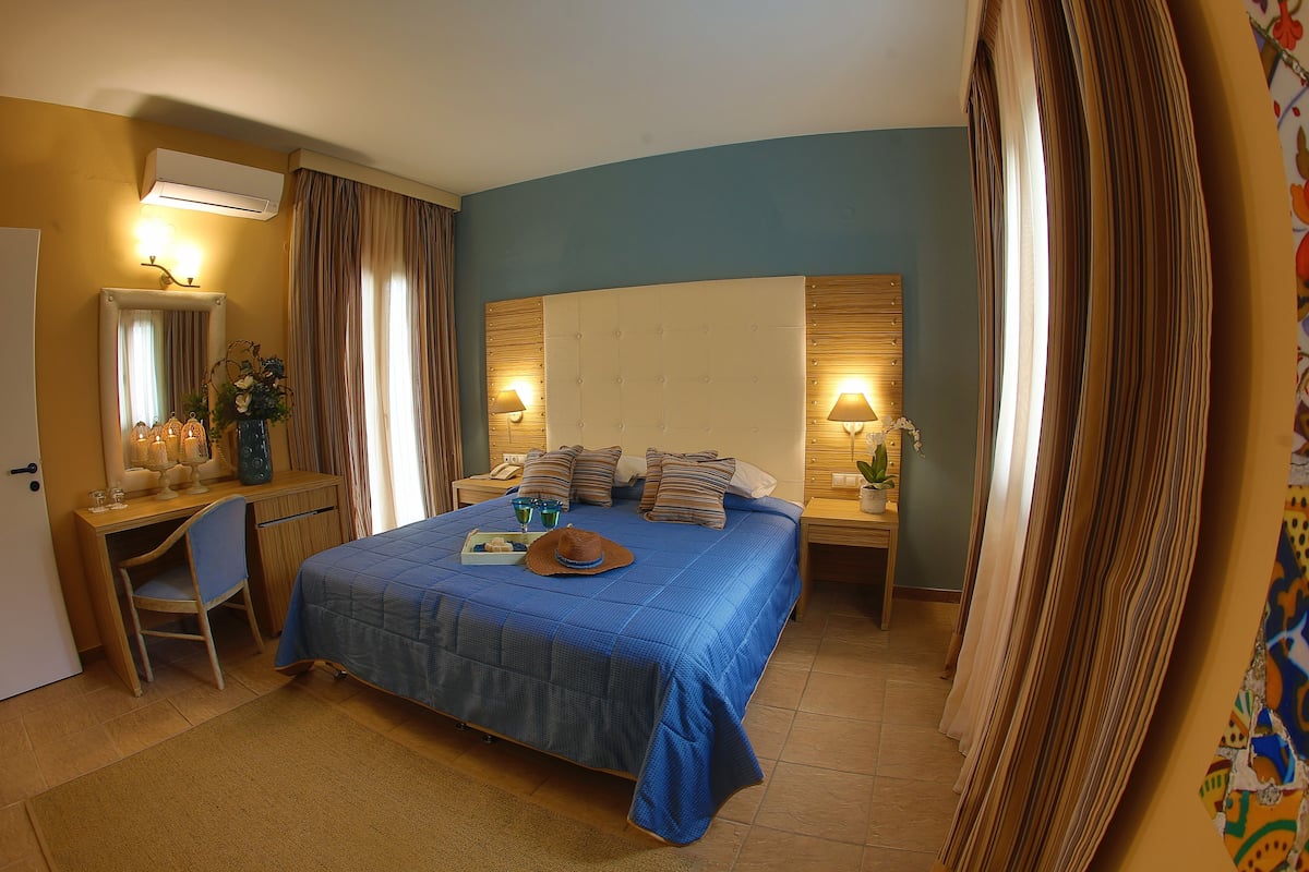 A hotel room with a large bed, a desk, a chair, and a mirror. There are two bedside tables with lamps and a small table with a vase and a hat.