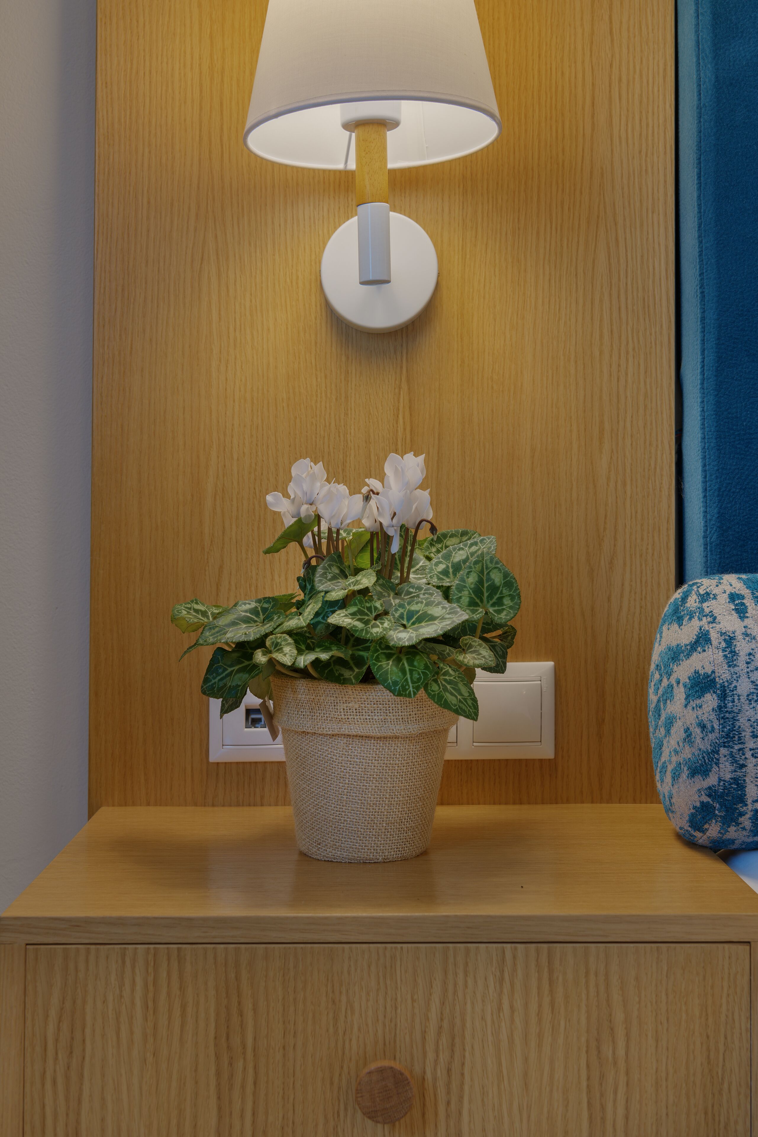 A wooden nightstand with a lamp, a potted plant with white flowers, and a decorative blue vase.
