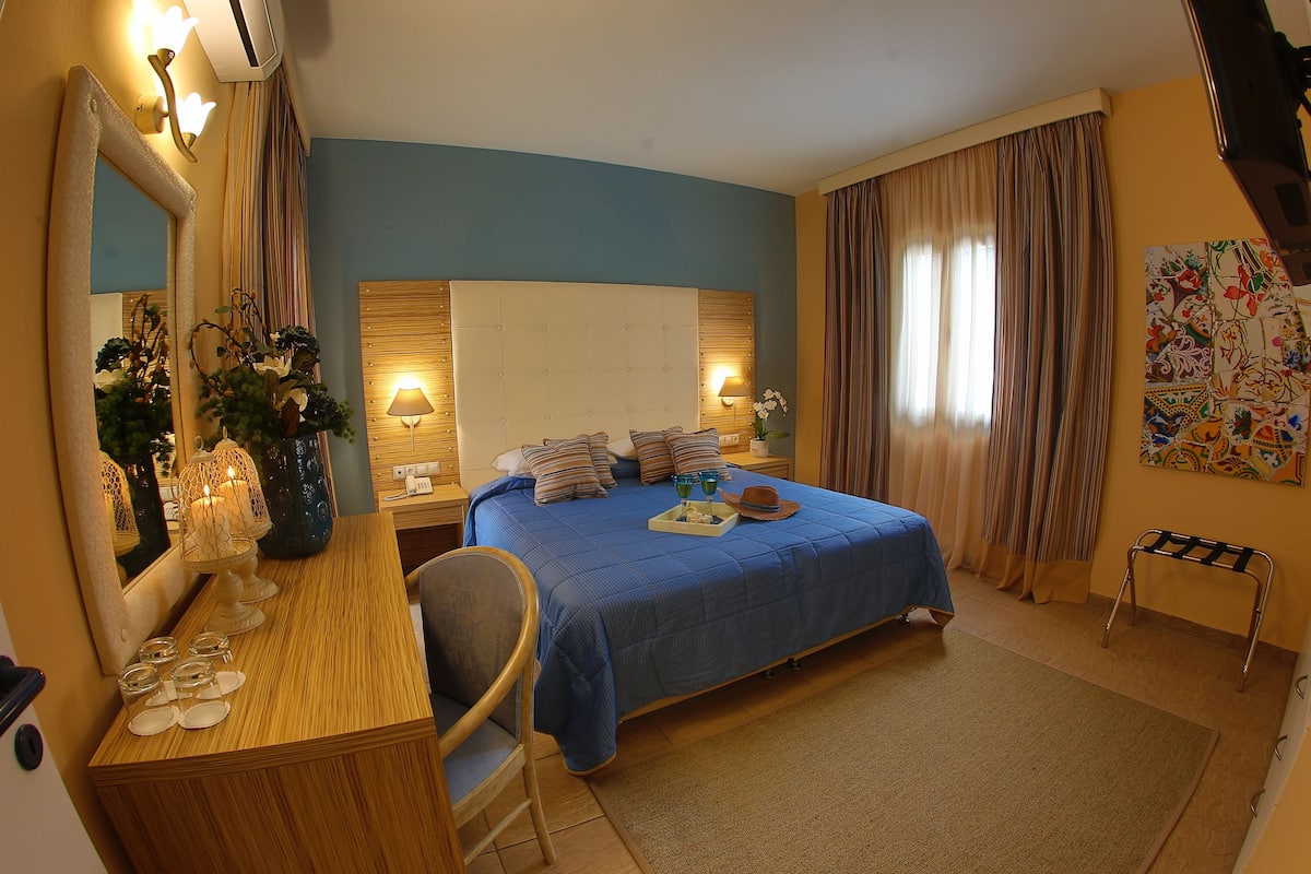 A hotel room with a large bed, a wooden desk, a chair, a mirror, and a painting on the wall.