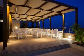 An outdoor dining area with white chairs and tables, covered by a roof, and overlooking a body of water at night.