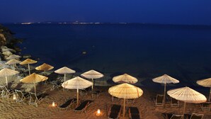 Una playa con filas de tumbonas y sombrillas de paja, un mar tranquilo y un paisaje urbano distante en la noche.
