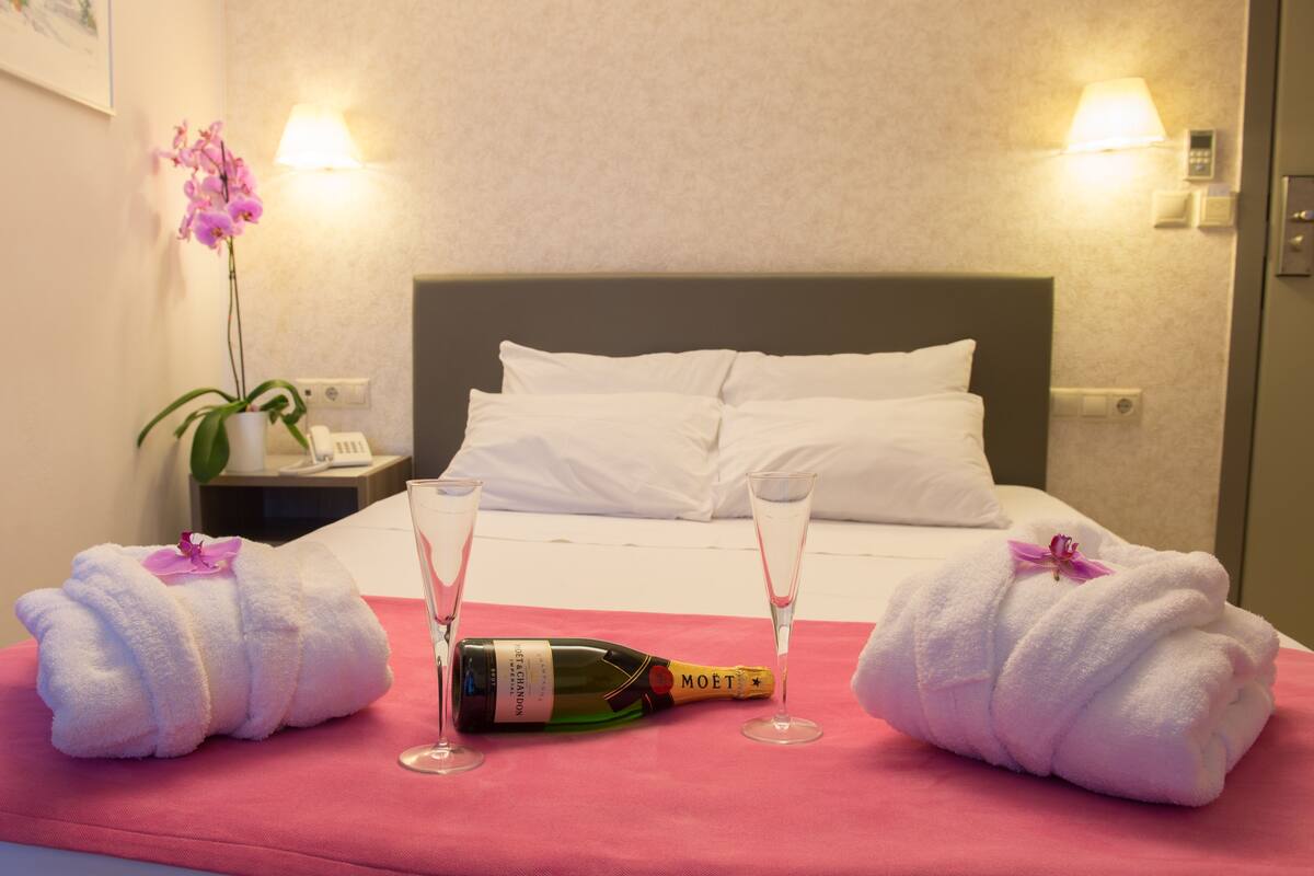 A hotel room with a bed, two towels, a bottle of champagne, and two glasses on a pink bedspread.