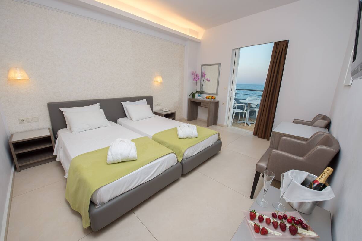 A hotel room with two beds, a desk with a bottle of champagne, and a balcony with a view of the sea.