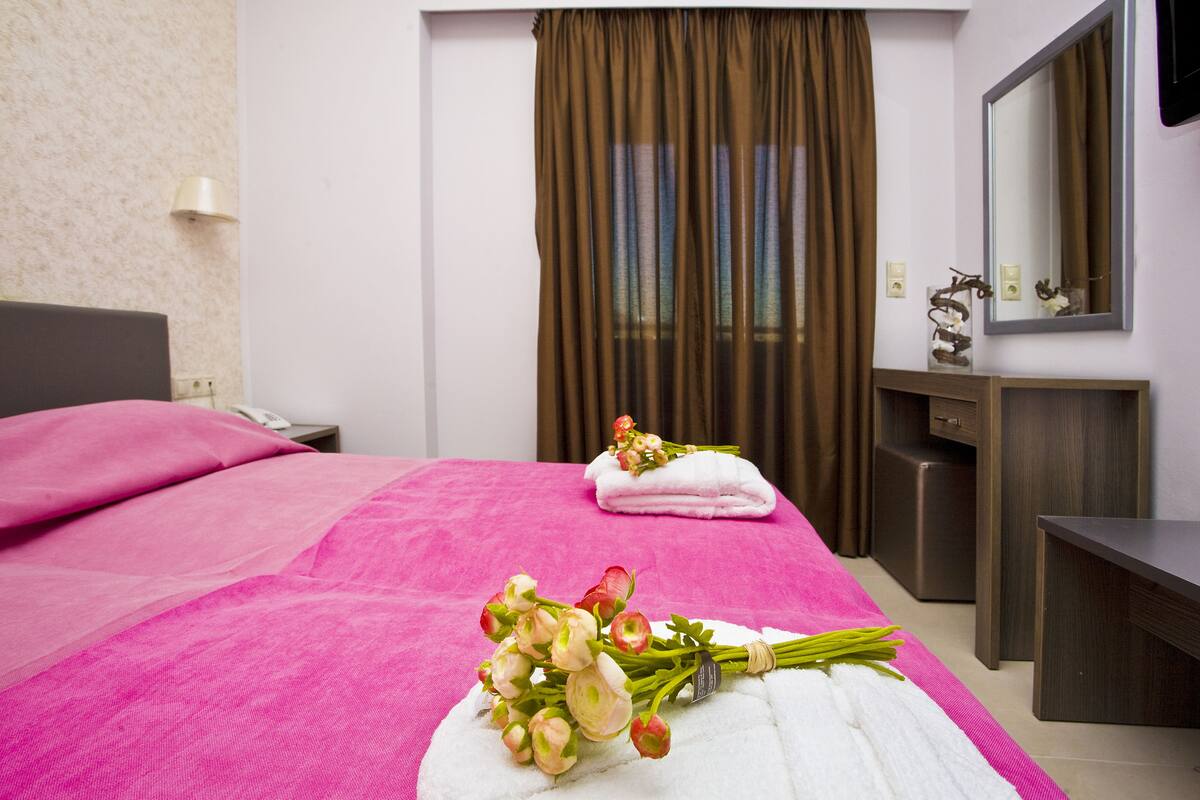 A hotel room with a bed covered in pink sheets, a white towel, and a bouquet of flowers on the bed. There is a wooden nightstand with a lamp and a phone, a wooden dresser, and a mirror on the wall.