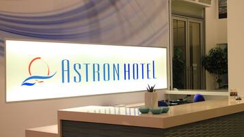 Una mesa de recepción con un letrero que dice "Astron Hotel" y un logotipo con un sol y una ola estilizados.