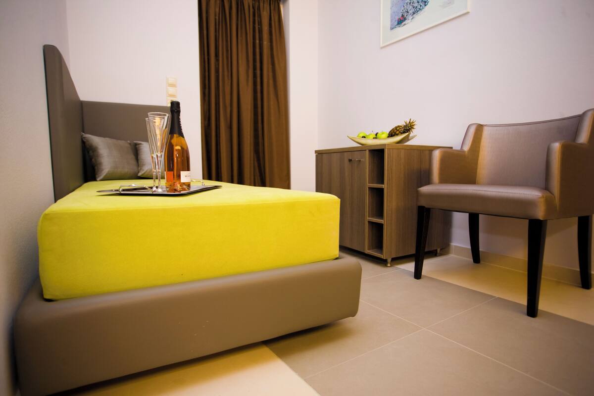 A modern hotel room with a bed, a chair, a small table with a bottle and glasses, and a cabinet with a fruit bowl.
