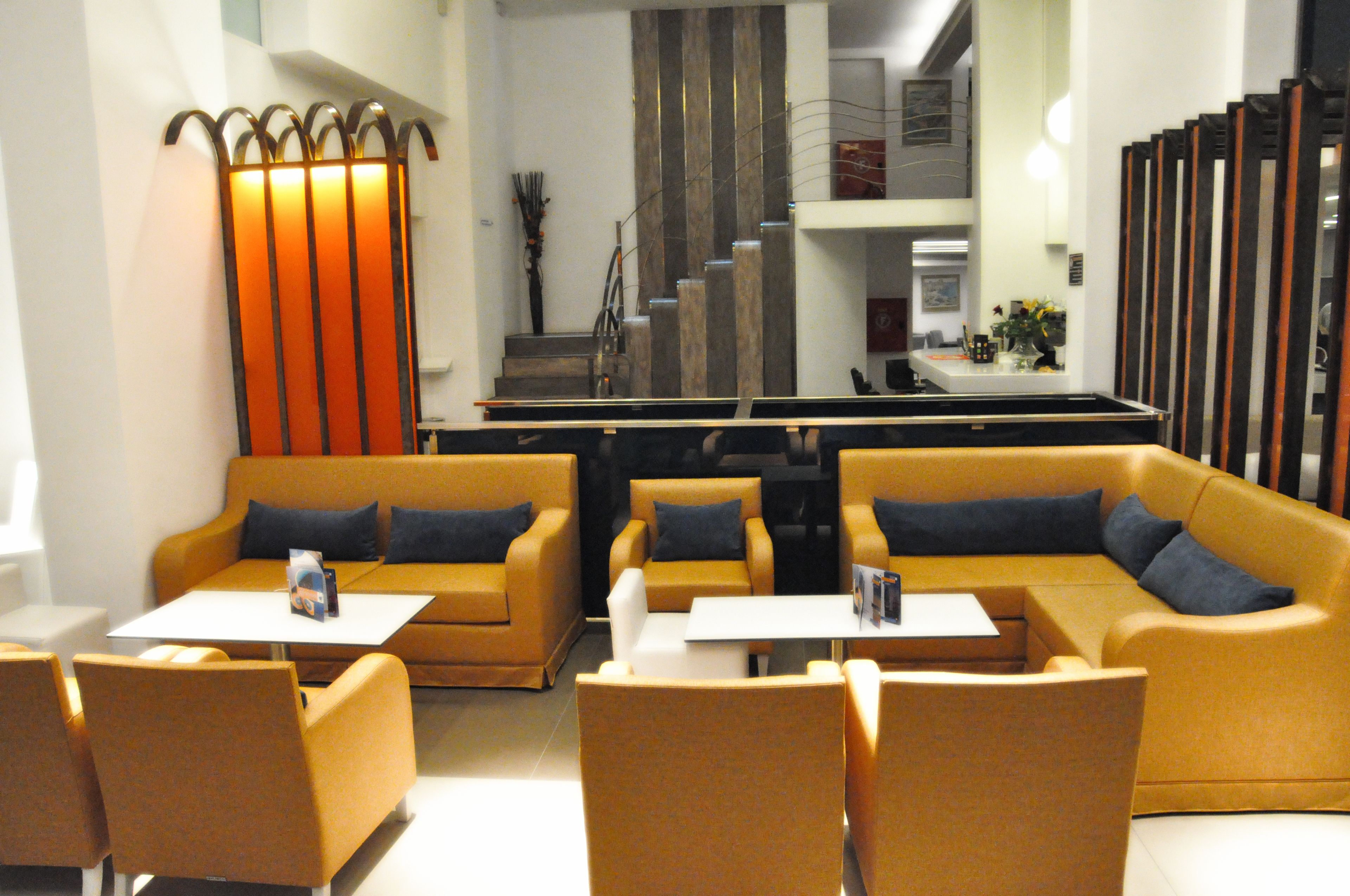 A modern hotel lobby with a seating area featuring sofas and armchairs, a reception desk, and a staircase in the background.