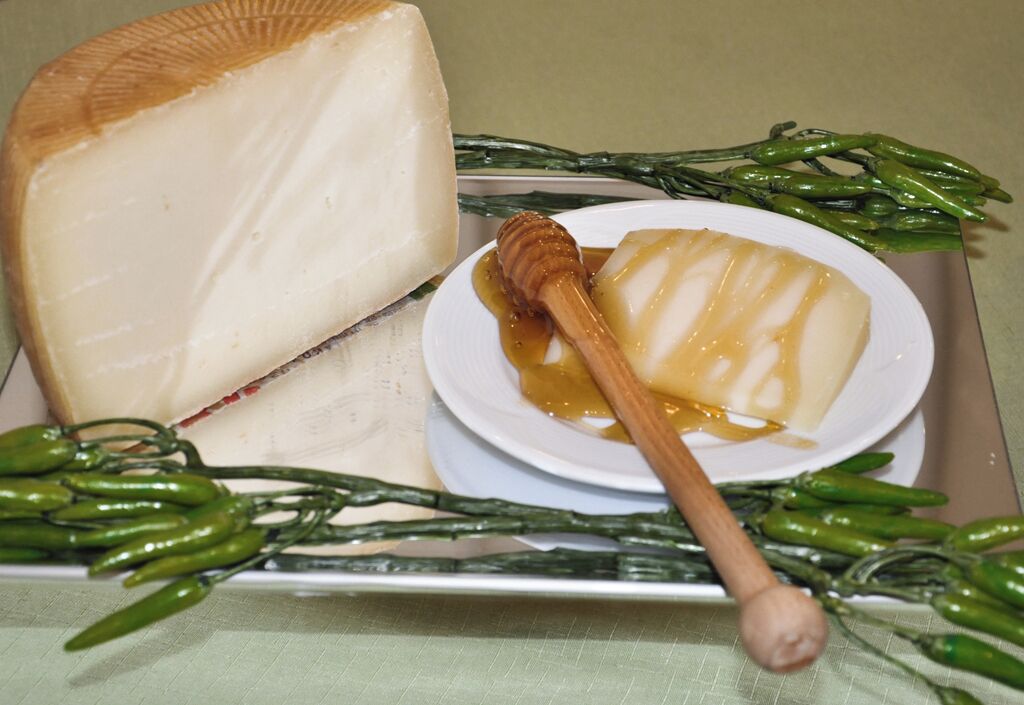 A large wheel of cheese, a bowl of honey, and green chili peppers on a tray.