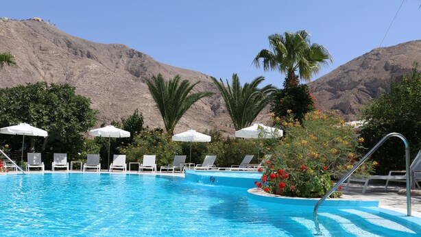 A swimming pool with lounge chairs and umbrellas, surrounded by palm trees and flowers, with mountains in the background. - Spiros & Hiroko Hotel - Adults Only (Santorini)