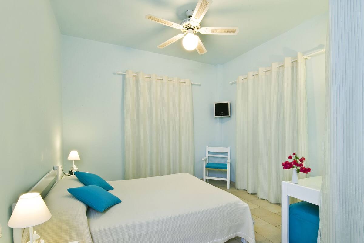 A hotel room with a bed, a chair, a small table with a vase, a ceiling fan, and curtains.