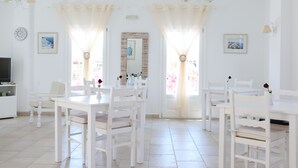 A dining area with white furniture, including tables and chairs, and a television on the left. - Spiros & Hiroko Hotel - Adults Only (Santorini)