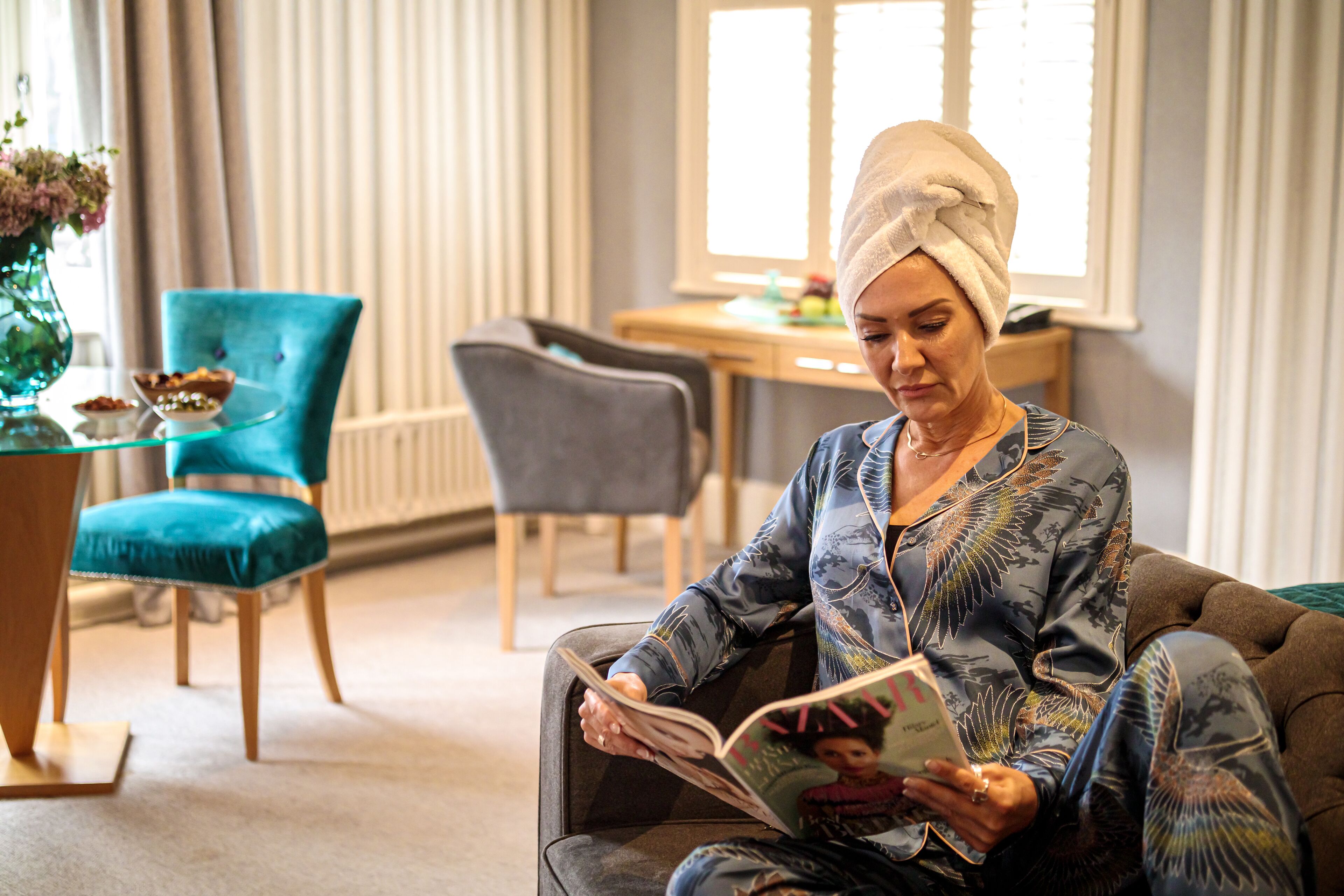 Een persoon zit op een bank, een boek lezend in een goed verlichte kamer, met een eettafel en stoelen op de achtergrond.