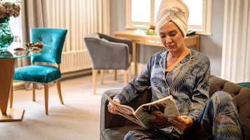 Een persoon zit op een bank, een boek lezend in een goed verlichte kamer, met een eettafel en stoelen op de achtergrond.