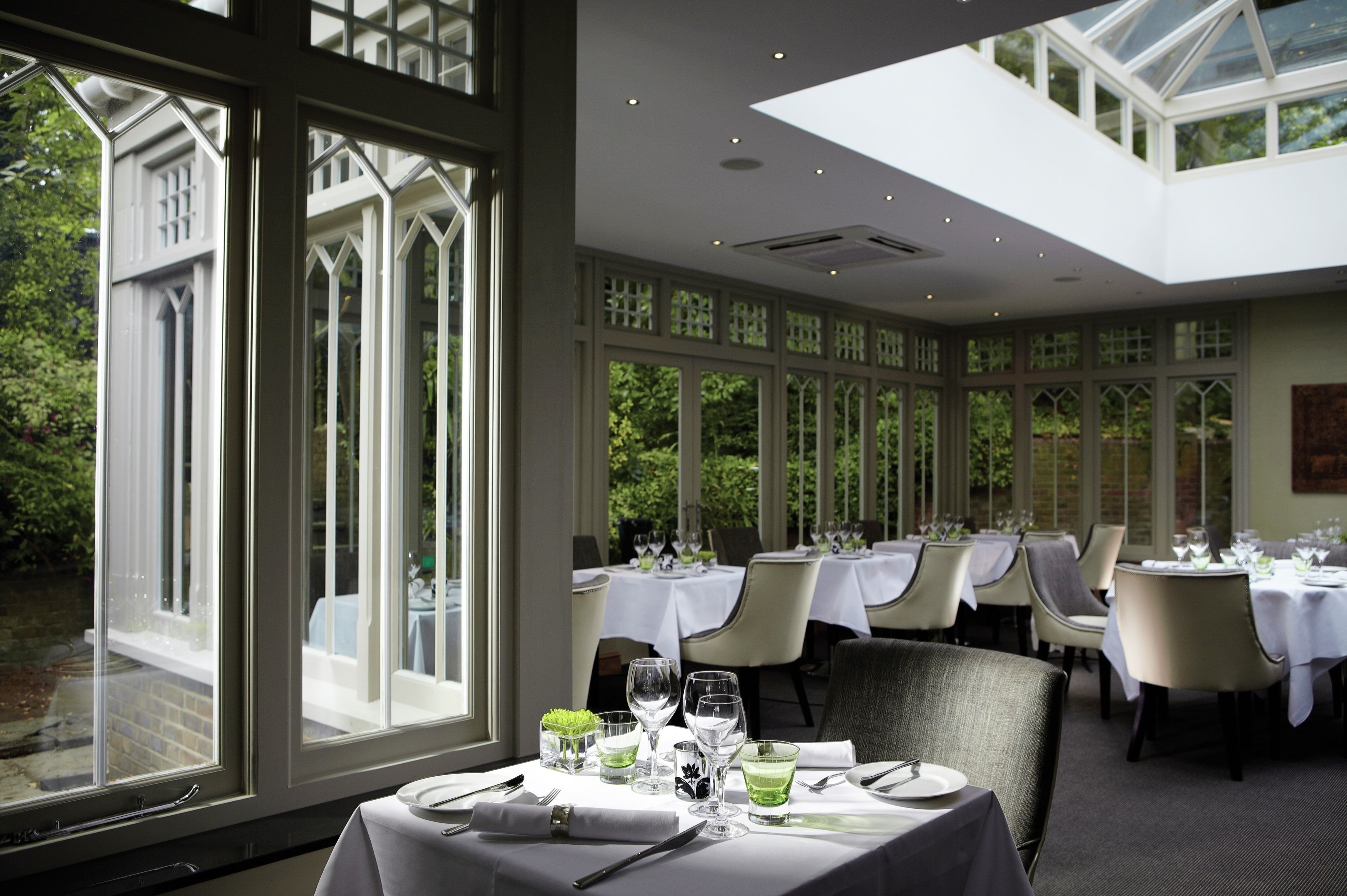 A restaurant with tables set for dining, large windows, and a view of greenery outside.