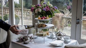 A table set for a tea service with a tiered tray holding sandwiches, scones, and pastries, a vase of flowers, and a bottle of champagne. - Rowhill Grange Hotel & Utopia Spa (Dartford)