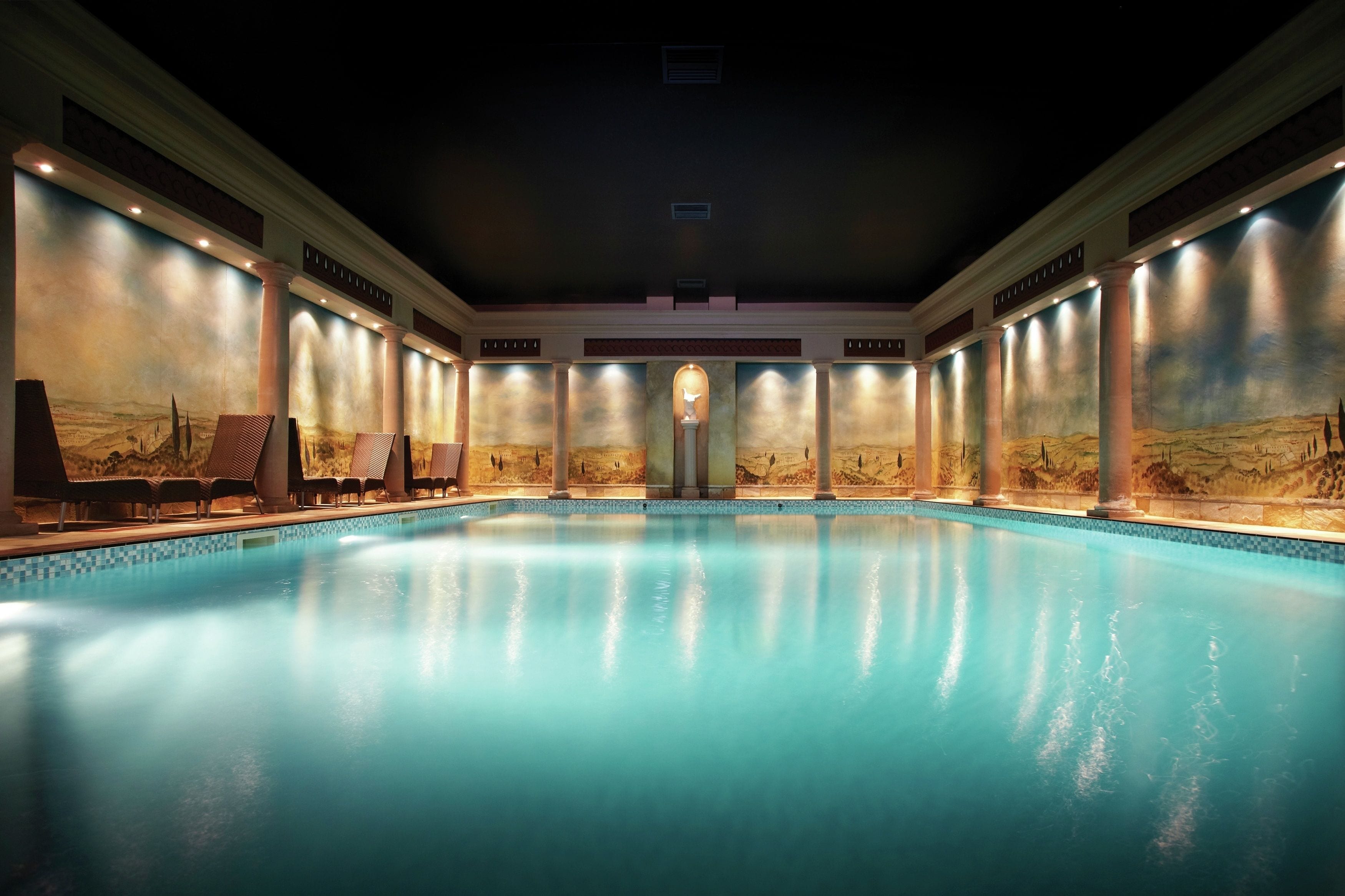 A large indoor swimming pool with a desert mural on the walls, multiple lounge chairs, and a central fountain.