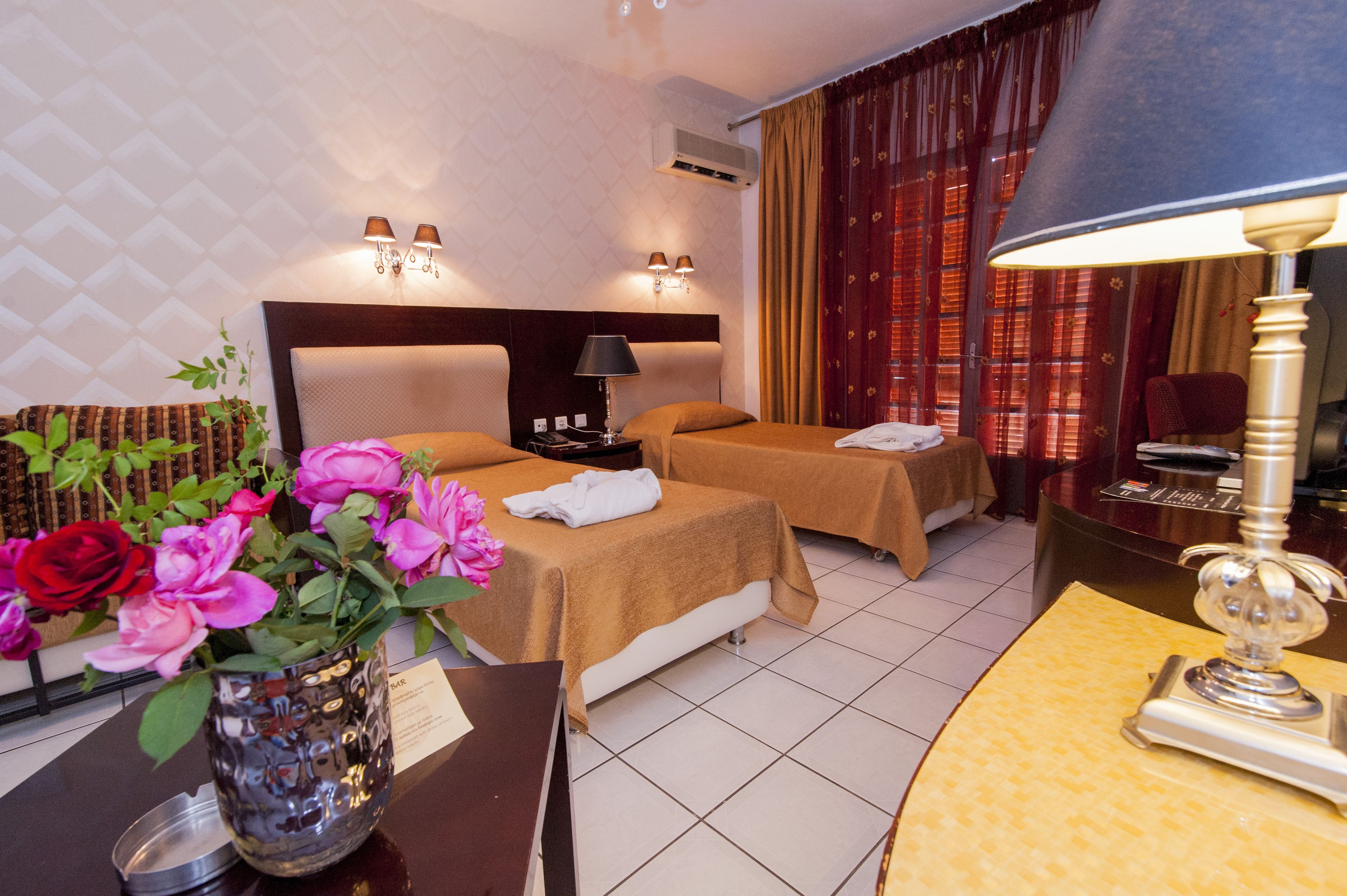 Una camera d'albergo con due letti, un comodino, una lampada, un vaso con fiori e una scrivania con lampada.