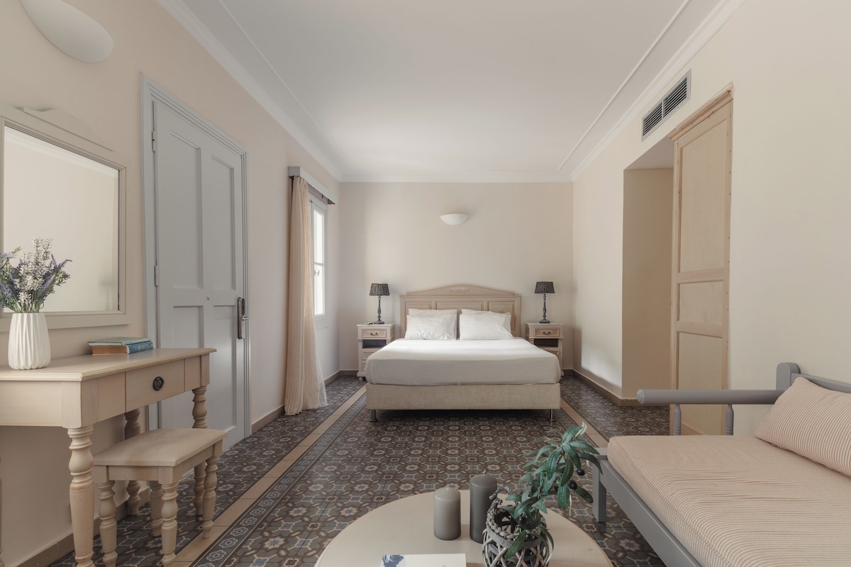 Habitación de hotel con cama, mesitas de noche, escritorio, silla, pequeña mesa con un florero y planta en maceta.