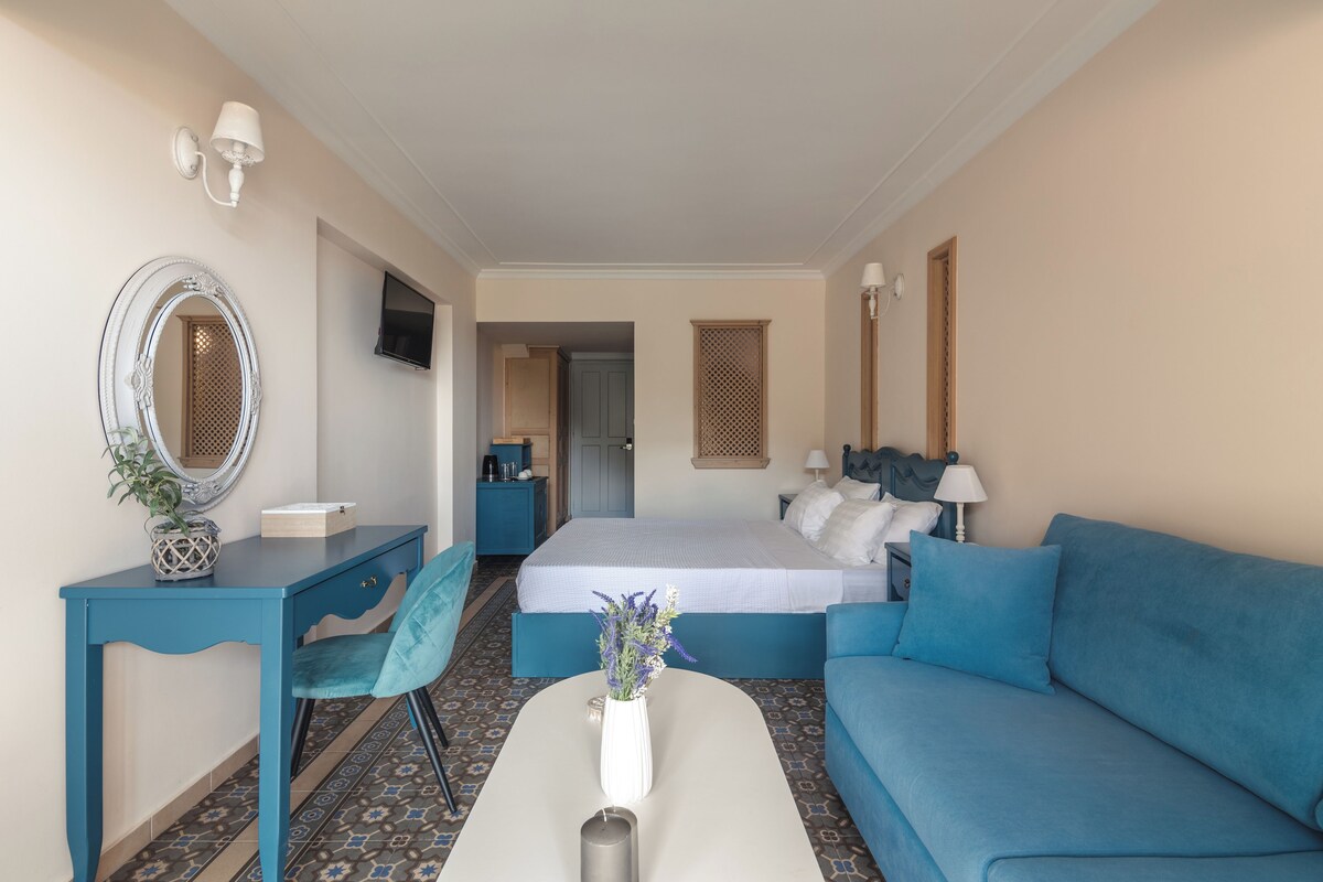Hotelkamer met een bed, een blauwe bank, een bureau met stoel, een spiegel en een kleine tafel met vaas.